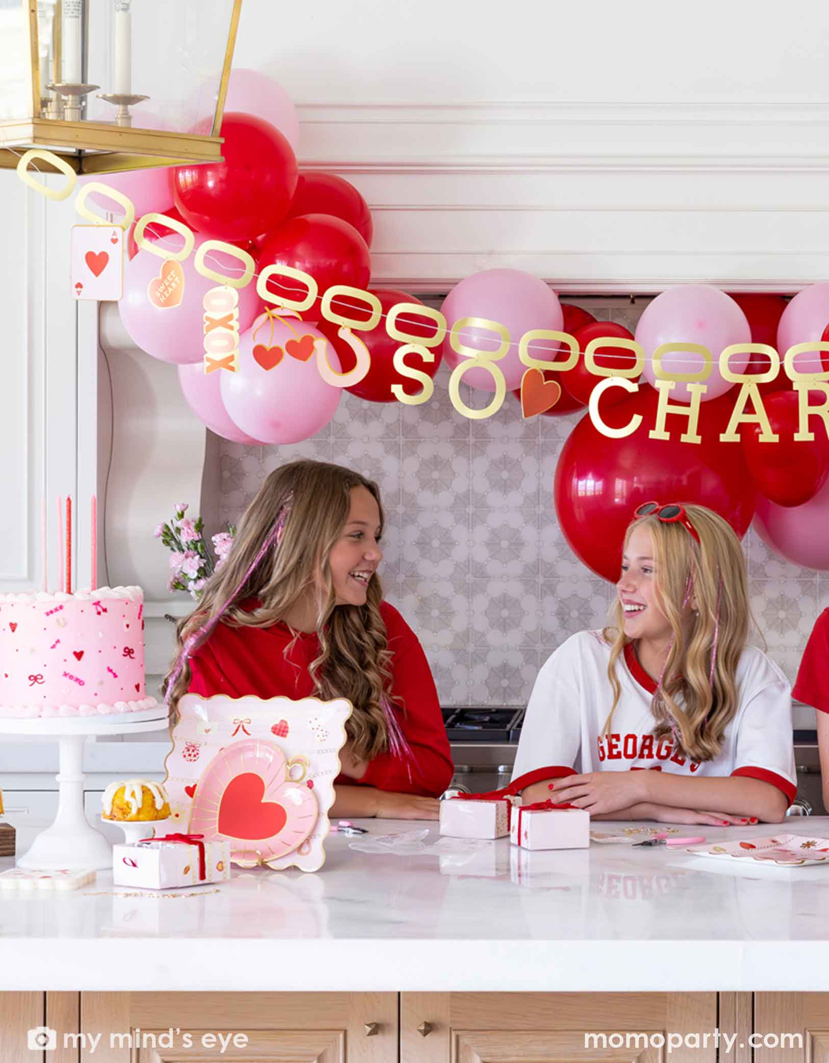 Two girls sitting at a Valentine's Day party table filled with heart themed party supplies from Momopartys including plates, napkins, and a cake. In the back there's a heart-themed balloon arch in red and pink and a gold foil party banner spells "So Charming". 