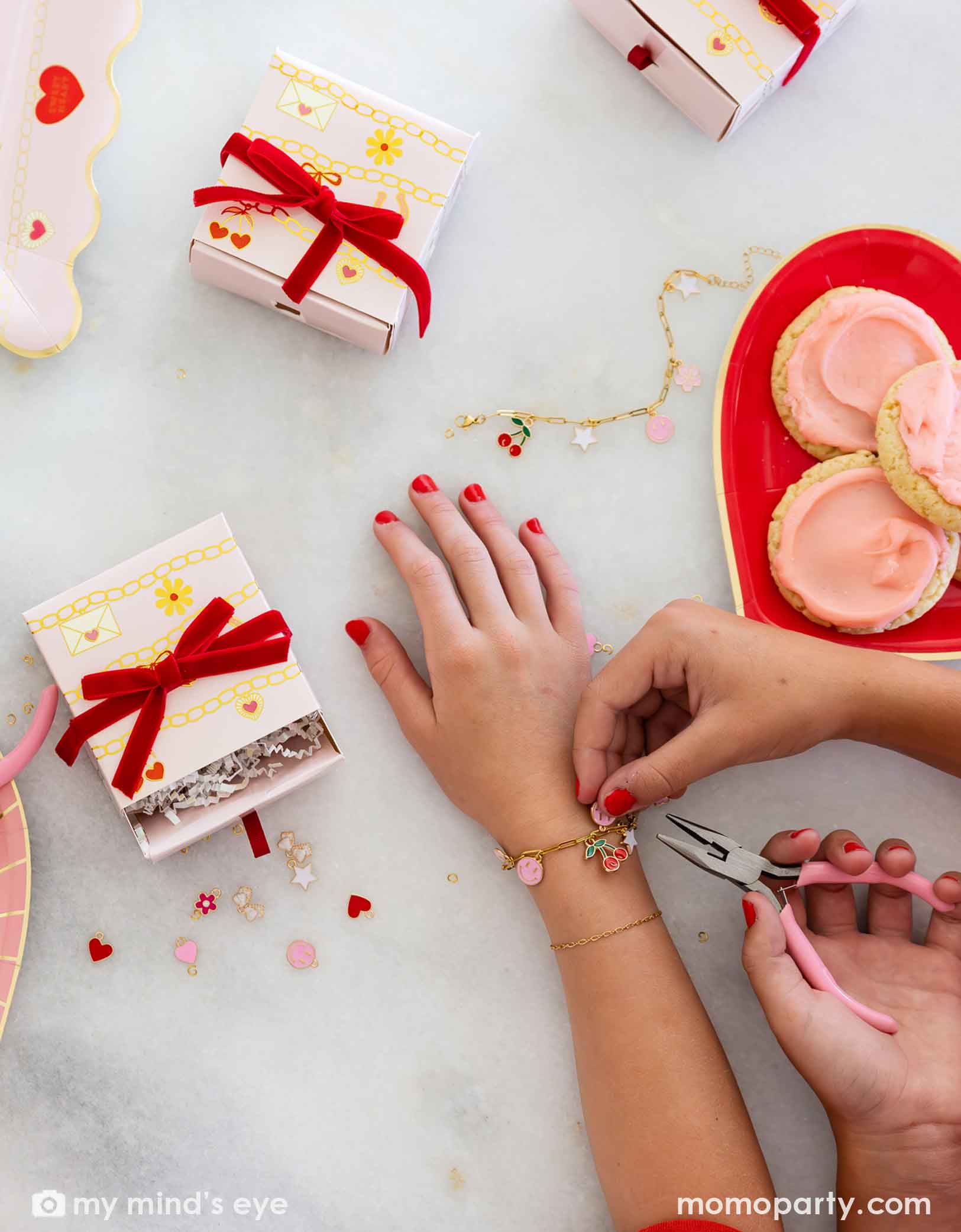 Children's hands with pink nail polish working on their charm bracelets with pink pliers, surrounded by small charms, gift boxes and Momopartys's Valentine's Day charm matching party tablewares including heart shaped plates and napkins on the table. 