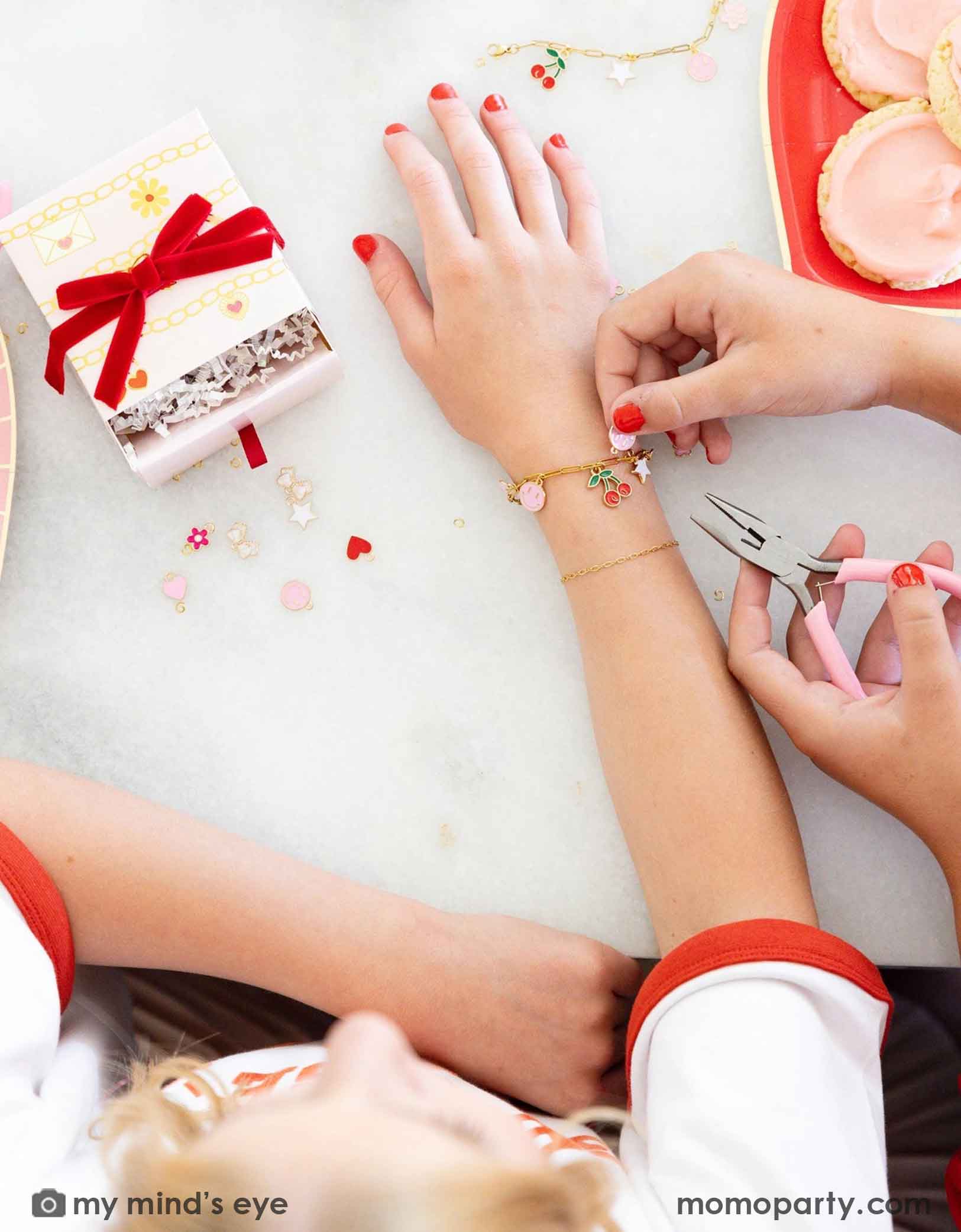 Children's hands with red nail polish working on their charm bracelets with pink pliers, surrounded by small charms, gift boxes and Momopartys's Valentine's Day charm matching party tablewares including heart shaped plates and napkins on the table. 