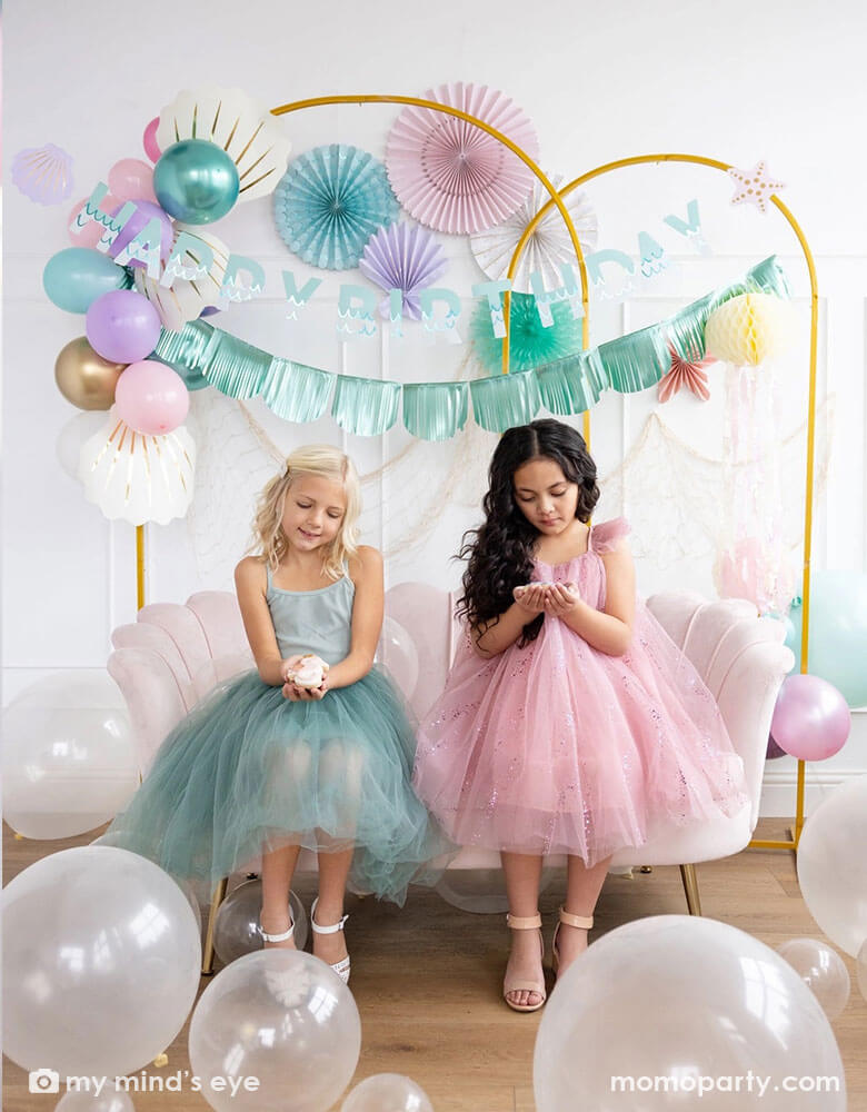 Two school-aged girls sitting on a shell shaped blush sofa in an enchanting kid's mermaid birthday party set up featuring mermaid inspired decorations and balloons from Momopartys including the Under the Sea paper fan set in pastel colors of light pink, aqua, teal, lilac and cream, Mermaid Happy Birthday Banner Set with aqua foil fringe banner and shell shaped paper decorations by My Mind's Eye on the wall. On the floor are some clear latex balloons in various sizes creating a whimsy water bubble scene.