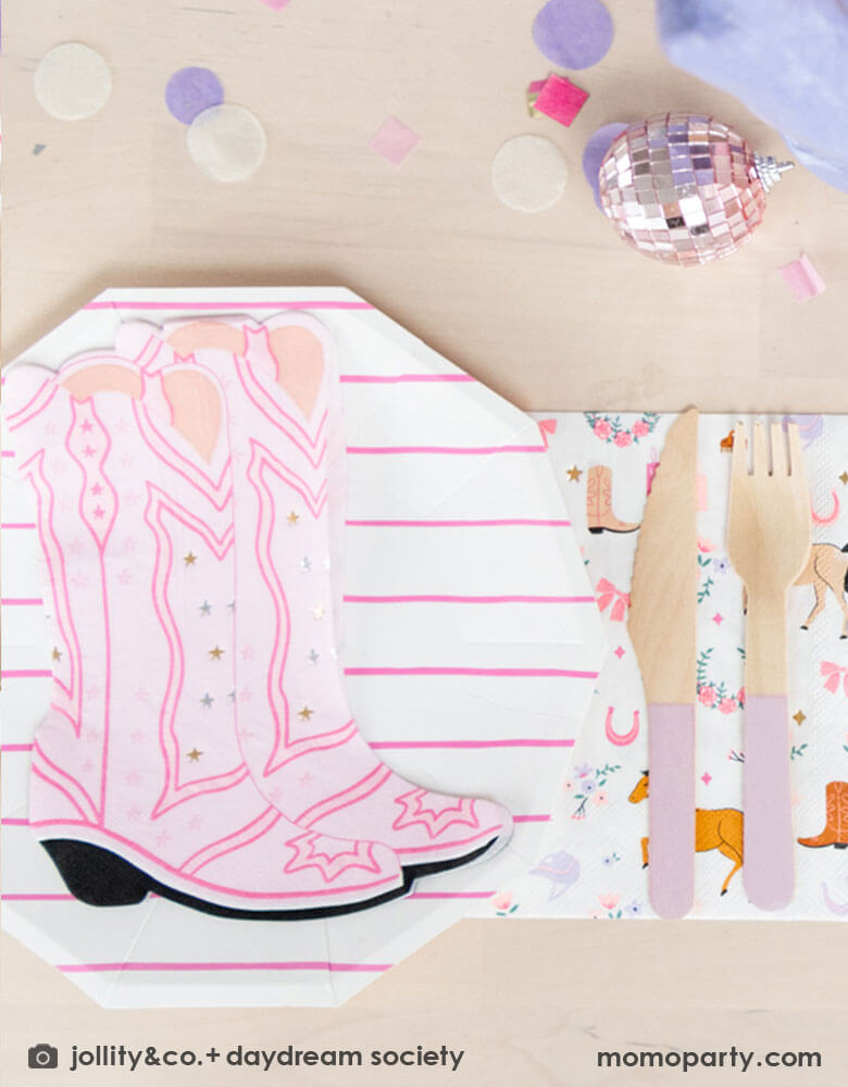 A girly cowgirl rodeo Western themed party table feature Momopartys's cowgirl boots shaped napkins, pink striped party plates, light pink wooden cutlery and cowgirl motif large napkins by Daydream Society. Around the tableware there are some lilac round confetti spread-out and a small pink disco ball decoration, making this an adorable inspiration for girl's First Rodeo first birthday celebration or an adorable cowgirl theme party for pony-loving girls!