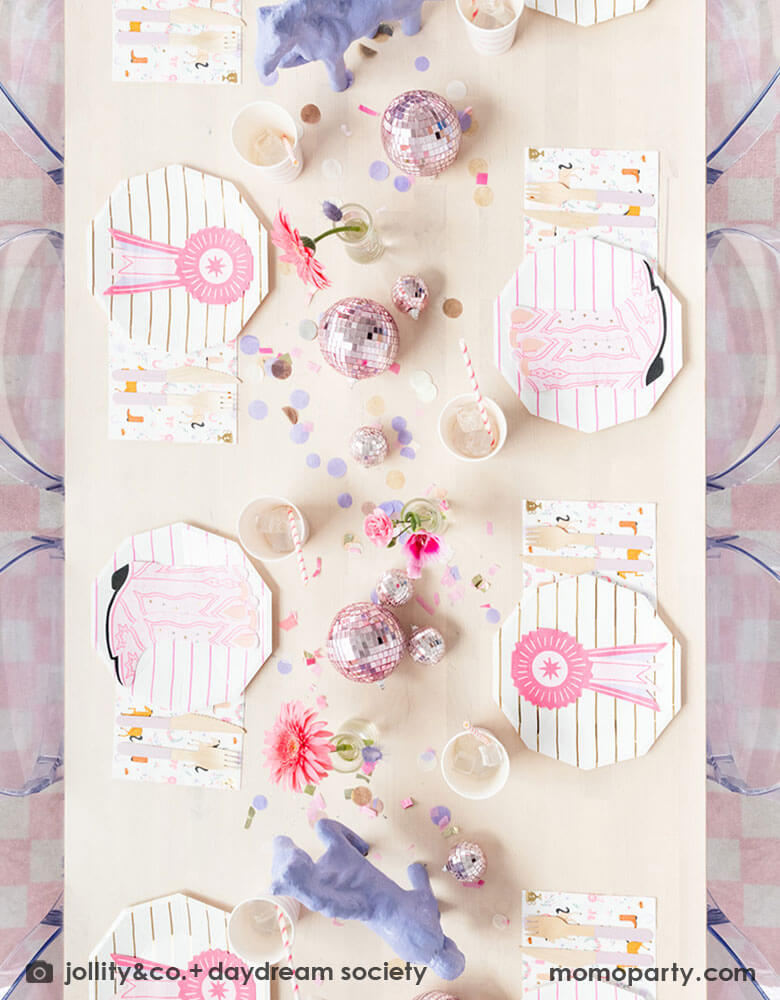 A table shot from the top of a girl's rodeo pony themed party table decoration features Momopartys's cowgirl themed party supplies by Daydream Society, including cowgirl boots shaped napkins, pink striped party plates, cups and cowgirl motif large napkins. In the middle of the party table, there are two purple pony figures as the centerpiece with some pink disco balls, making this an adorable inspiration for girl's First Rodeo first birthday celebration or a cowgirl theme party for pony-loving girls!