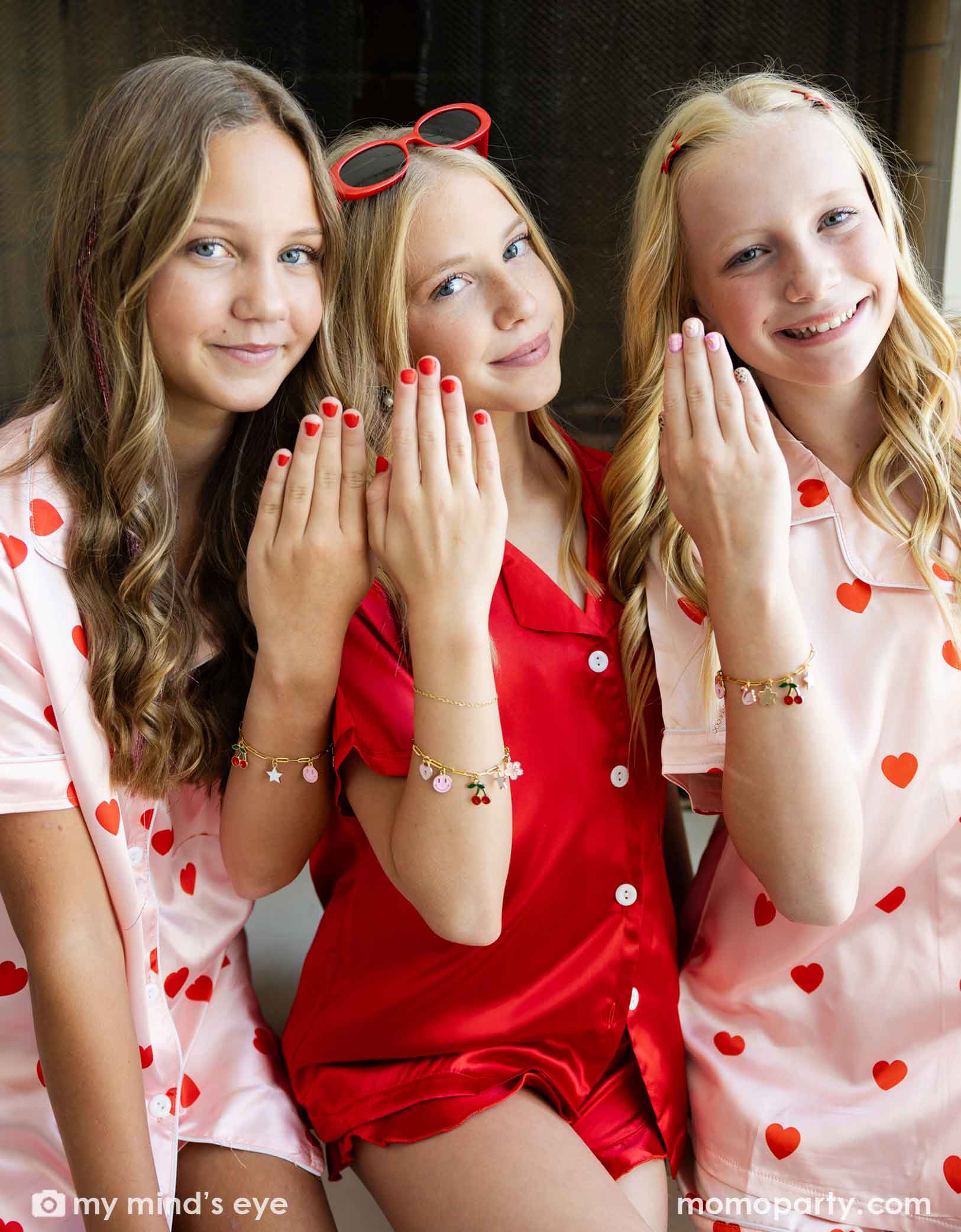 Three young girls in Valentine's Day pajamas with heart patterns showing their hands with red and pink polish and the charm bracelets from Momopartys posing together.