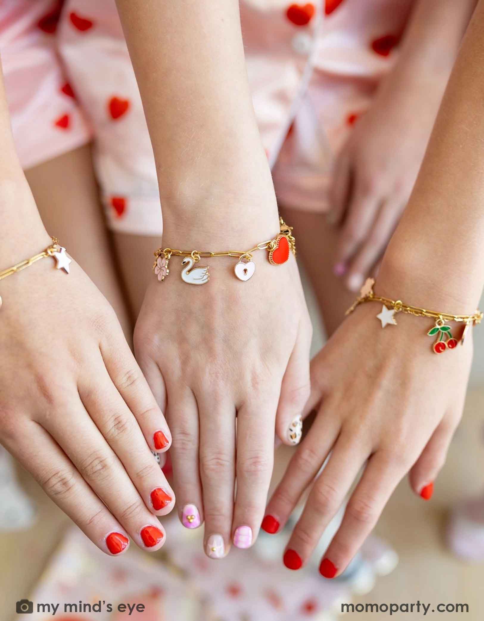 Three girls' hands with red nail polish showing their gold bracelets with various charms from Momopartys against a blurred background at a Valentine's Day themed sleep over party celebration.