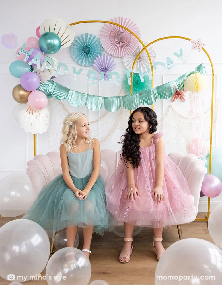 Two school-aged girls sitting on a shell shaped blush sofa in an enchanting kid's mermaid birthday party set up featuring mermaid inspired decorations and balloons from Momopartys including the Under the Sea paper fan set in pastel colors of light pink, aqua, teal, lilac and cream, Mermaid Happy Birthday Banner Set with aqua foil fringe banner and shell shaped paper decorations by My Mind's Eye on the wall. On the floor are some clear latex balloons in various sizes creating a whimsy water bubble scene.