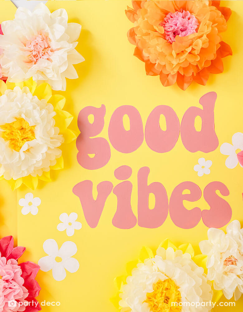 A yellow party backdrop wall with with GOOD VIBES in retro font adorn with cream, pink and orange tissue paper pom pom flower decorations from Momopartys.