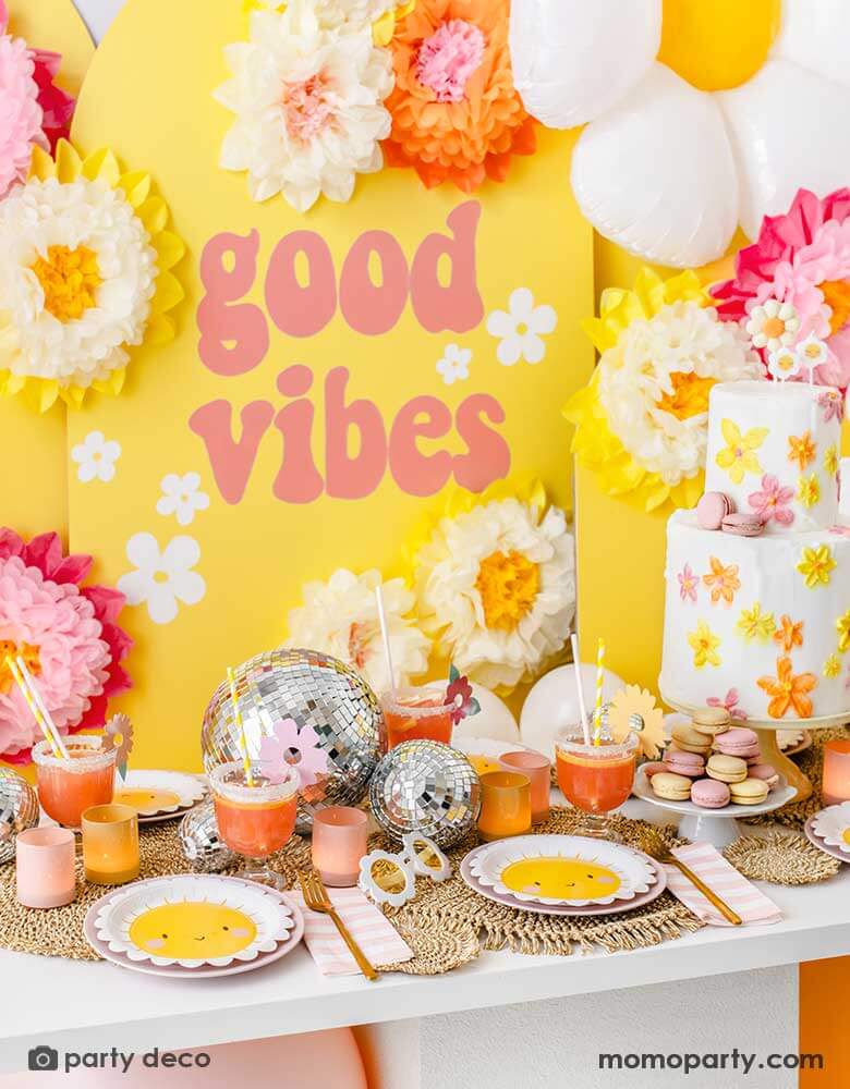 A "Good Vibes" themed party featuring Momopartys's scallop edged sun plates, pink and white striped napkins and sunflower shaped sunglasses. On the table is a two-tier butter cream birthday cake decorated with bright flowers in orange, yellow and peach colors, along with some color-matching macarons and drinks and fun disco balls as the centerpiece. In the back is a yellow backdrop with GOOD VIBES in retro font adorn with tissue paper pom pom flower decorations.