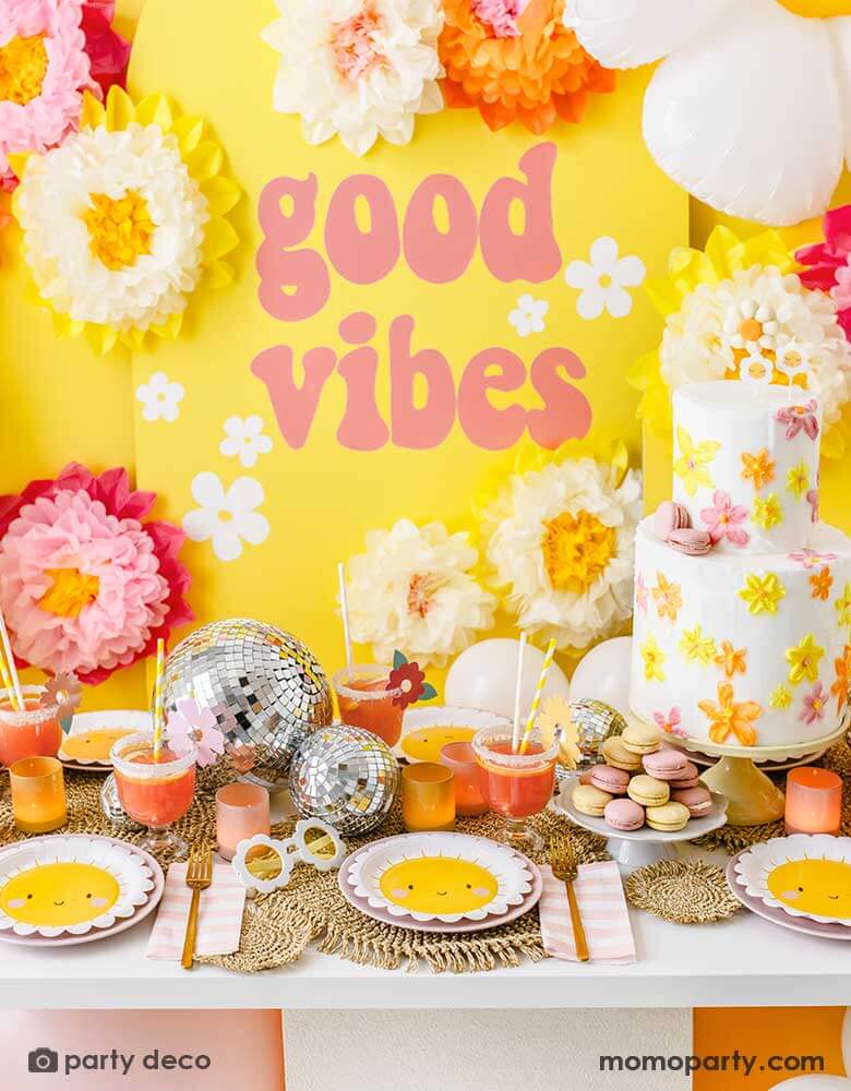 A "Good Vibes" themed party featuring Momopartys's scallop edged sun plates, pink and white striped napkins and sunflower shaped sunglasses. On the table is a two-tier butter cream birthday cake decorated with bright flowers in orange, yellow and peach colors, along with some color-matching macarons and drinks and fun disco balls as the centerpiece. In the back is a yellow backdrop with GOOD VIBES in retro font adorn with tissue paper pom pom flower decorations.