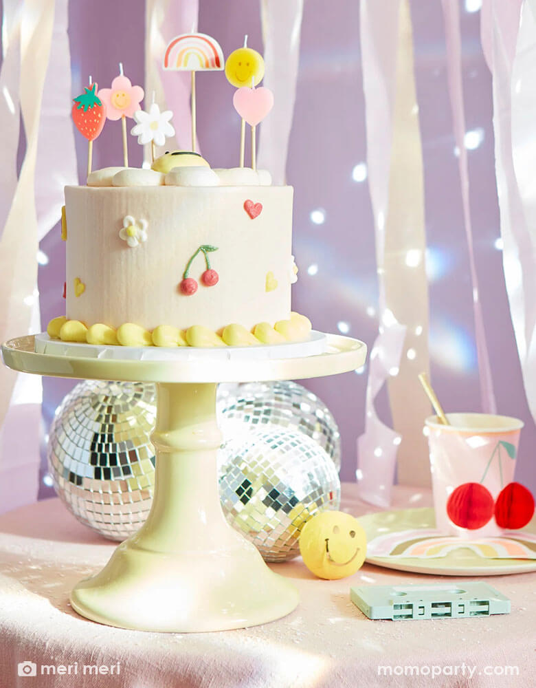 A festive retro themed birthday party table featuring a pale buttercream birthday cake topped with Momopartys's Happy Face Icons candle which includes a strawberry, a rainbow, a smiley face, a heart, and a couple of flowers. On the table there are a few disco balls and cassette tapes as the centerpiece, along with Meri Meri's happy faces cheery honeycomb paper cup and a rainbow napkin, with pastel streamers in the back, makes this a great inspo for a fun 90s' retro themed party.