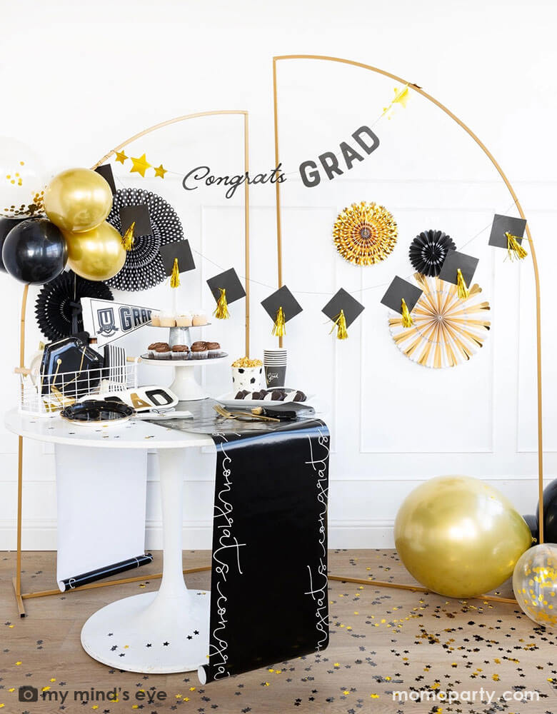 A graduation party featuring Momopartys's graduation party supplies by My Mind's Eye including U did it Grad felt party pennant, black striped dinner napkins, graduation cap shaped plates. On the table there are some cupcakes, cookies and popcorn on a black congratulation paper table runner and some black grad party cups and grad cap shaped napkins. In the back there are a gold backdrop frame adorned with some black and gold balloons and party fans, along with a Congrats Grad and grad cap tassels banner.