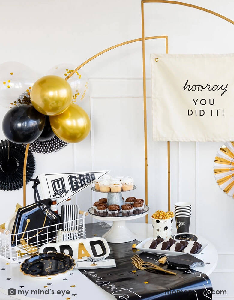 A graduation party featuring Momopartys's graduation party supplies by My Mind's Eye including U did it Grad felt party pennant, class of 2024 black striped dinner napkins, graduation cap shaped plates. On the table there are some cupcakes, cookies and popcorn on a black congratulation paper table runner and some black grad party cups and grad cap shaped napkins.  In the back there are a gold backdrop frame adorned with some black and gold balloons and party fans, along with a "Hooray You Did It!" banner.