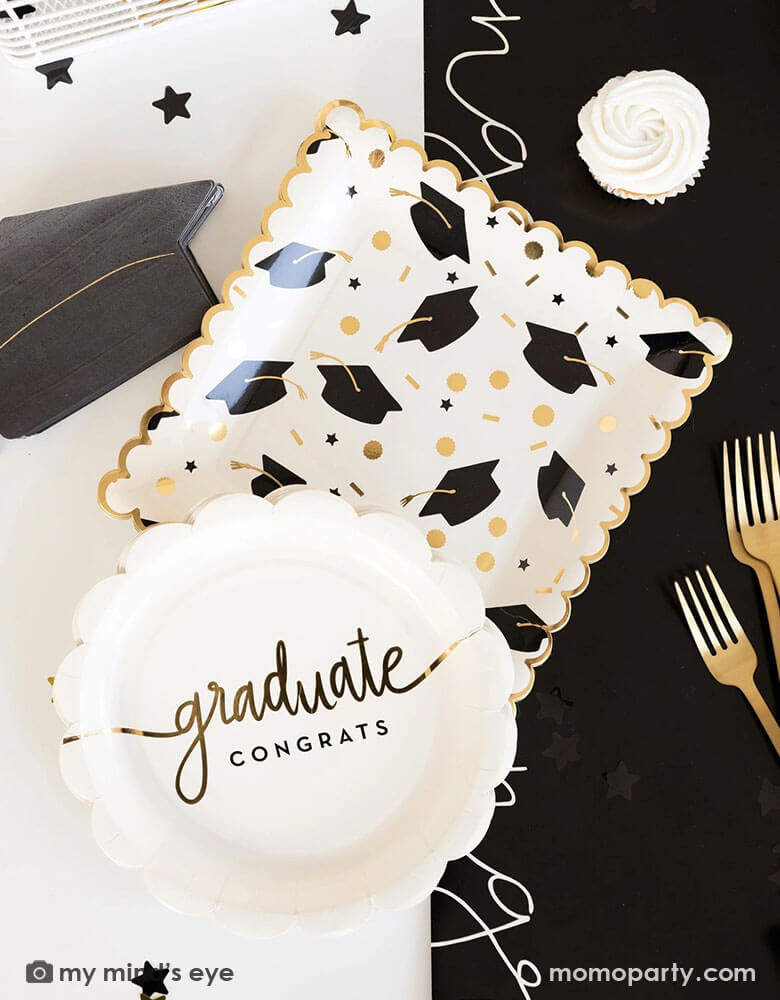 A graduation party table features Momopartys's 9" x 9" black and gold cap scatter paper plates, scalloped congrats graduate paper plates, grad cap shaped napkins and congratulation black paper runner along with some black and gold star shaped confetti by My Mind's Eye. Along with some cupcakes and gold utensils making this a great inspo for a modern and stylish graduation party tablescape.