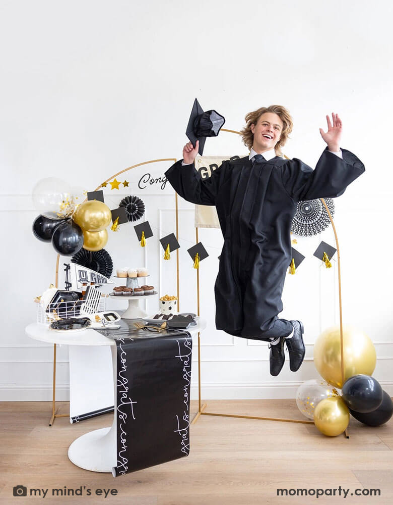 A graduate in his robe and cap jumping in a graduate party set up featuring Momopartys's graduation supplies including U did it Grad felt party pennant, black striped dinner napkins, graduation cap shaped plates. On the table there are some cupcakes, cookies and popcorn on a black congratulation paper table runner and some black grad party cups and grad cap shaped napkins. In the back there are a gold backdrop frame adorned with some black and gold balloons and party fans, along with a Congrats Grad banner.