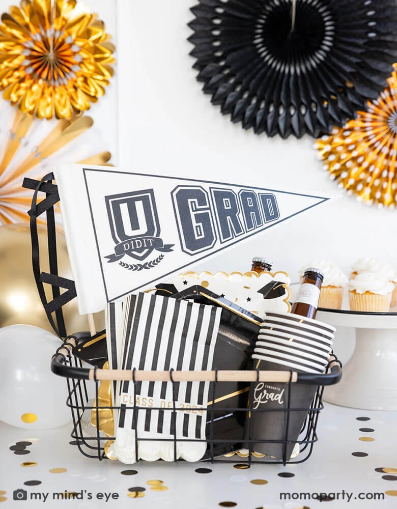 A graduation party set up featuring an occasion bin filled with Momopartys's graduation party supplies by My Mind's Eye including U did it Grad felt party pennant, class of 2024 black striped dinner napkins, graduation cap shaped plates, 12 oz grad party cups and grad cap scattered paper plates. In the back there are black and gold party fans hung on the wall and some cupcakes and graduation confetti on the table. Making this a perfect inspo for modern and stylish graduation party set up.