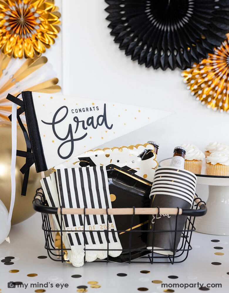 A graduation party set up featuring an occasion bin filled with Momopartys's graduation party supplies by My Mind's Eye including Congrats Grad felt party pennant, class of 2024 black striped dinner napkins, graduation cap shaped plates, 12 oz grad party cups and grad cap scattered paper plates. In the back there are black and gold party fans hung on the wall and some cupcakes and graduation confetti on the table. Making this a perfect inspo for modern and stylish graduation party set up.
