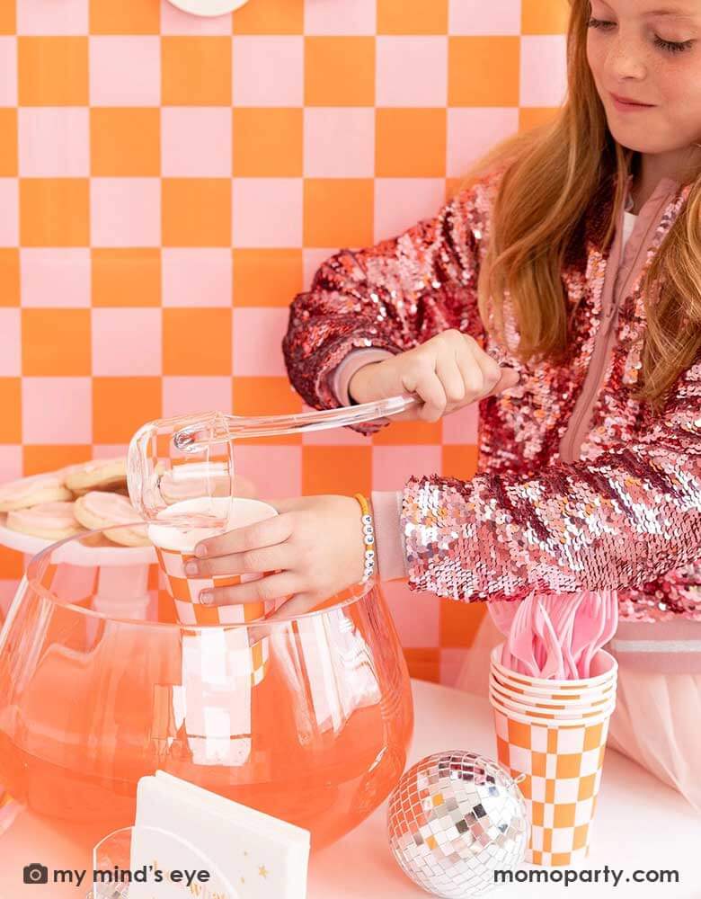 A girl in her pink sequin jacket stands in front of a wall covered with an orange and pink wallpaper. She's pouring some punch into Momopartys's 10oz orange and pink checkered party cup, next to the punch bowl is a disco ball decoration and more checkered cups with pink utensils for a groovy themed Halloween party in this spooky season.