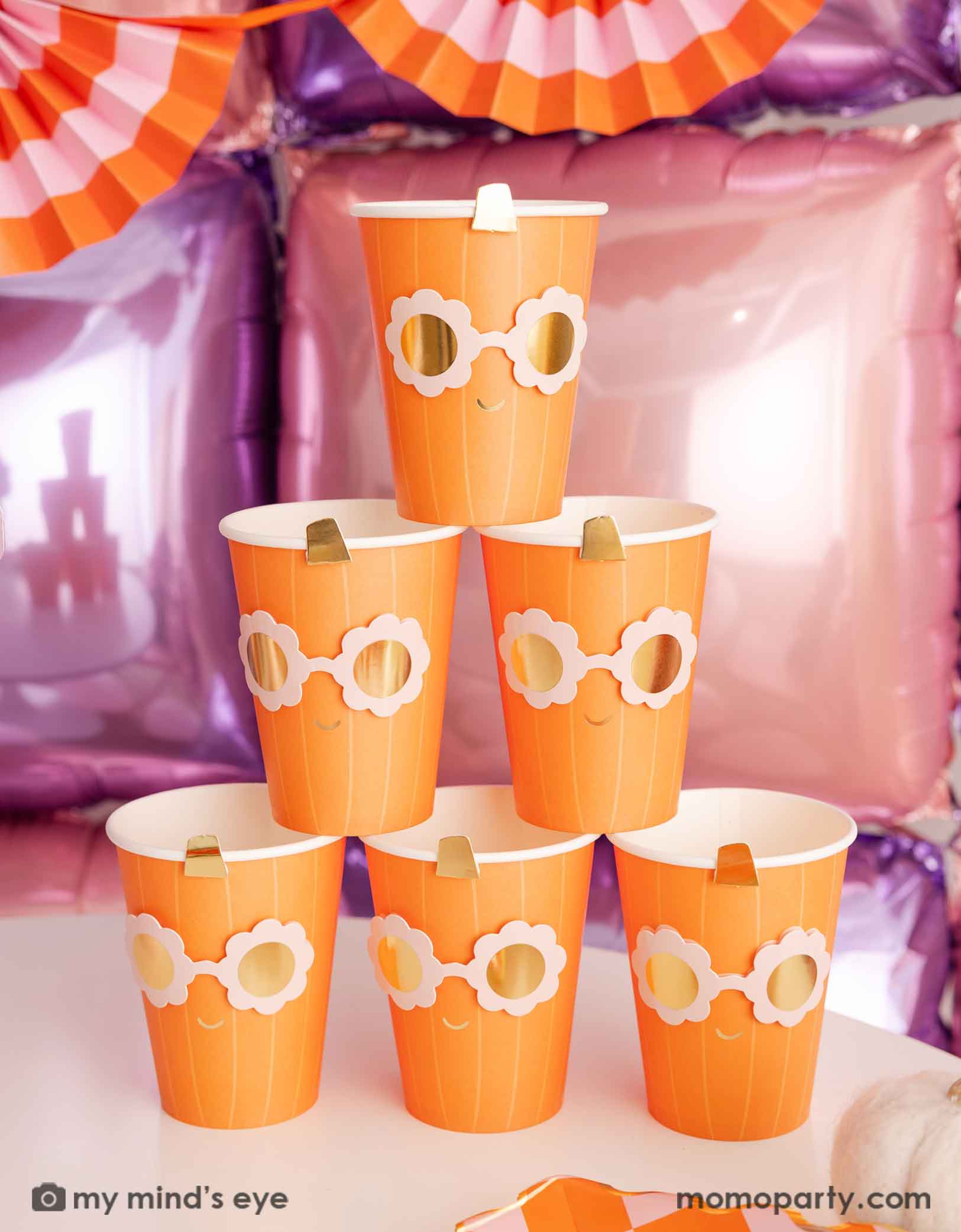 A stack of the orange pumpkin shaped paper party cups with daisy-shaped sunglasses from Momopartys on a Halloween inspired background with a foil balloon wall in pink and lilac and an orange and pink bunting garland. 