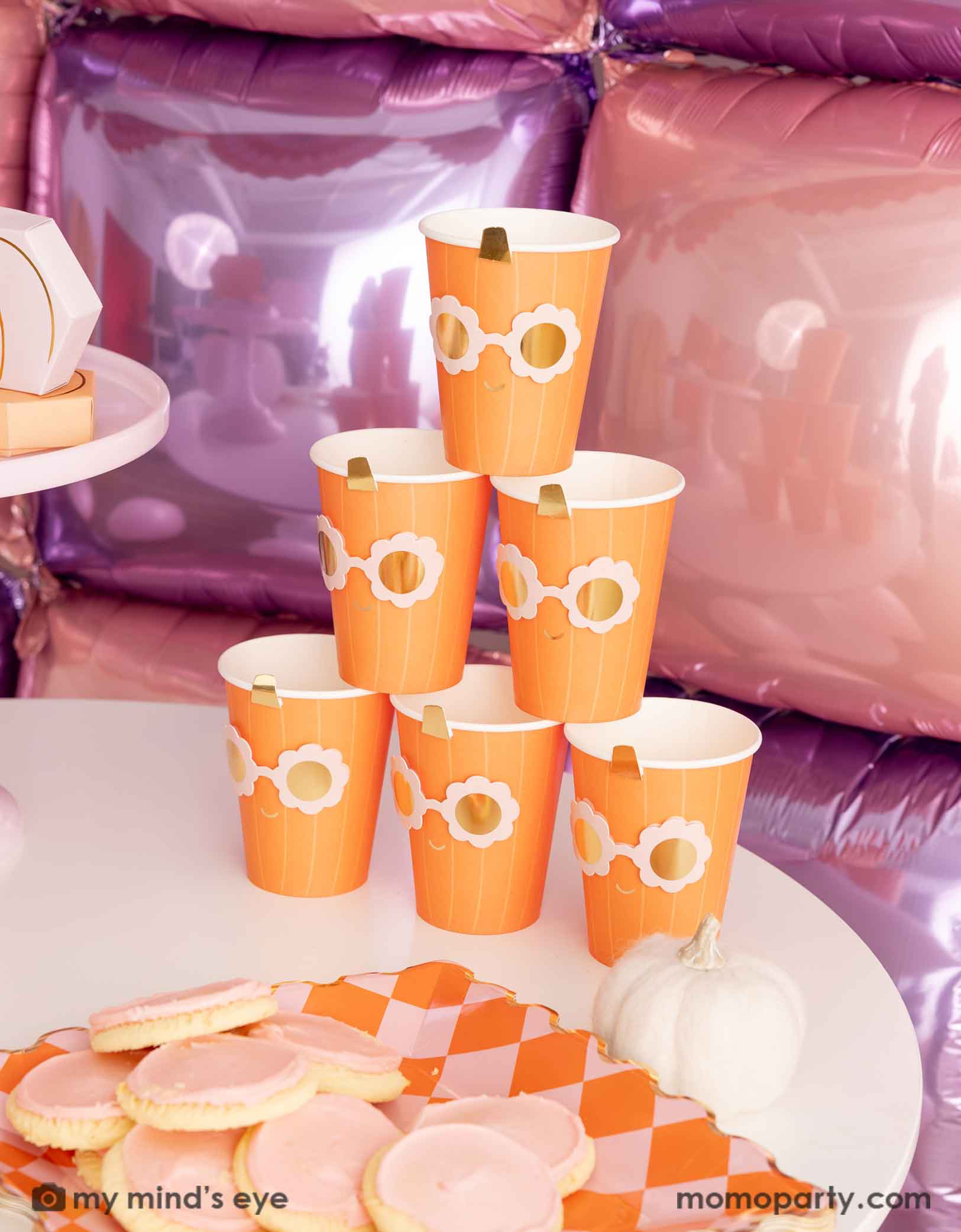 A stack of the orange pumpkin shaped paper party cups with daisy-shaped sunglasses from Momopartys on a Halloween inspired background with a foil balloon wall in pink and lilac. On the table there's  a orange and pink checkered dinner plate filled with pink sugar coated cookies for a party.