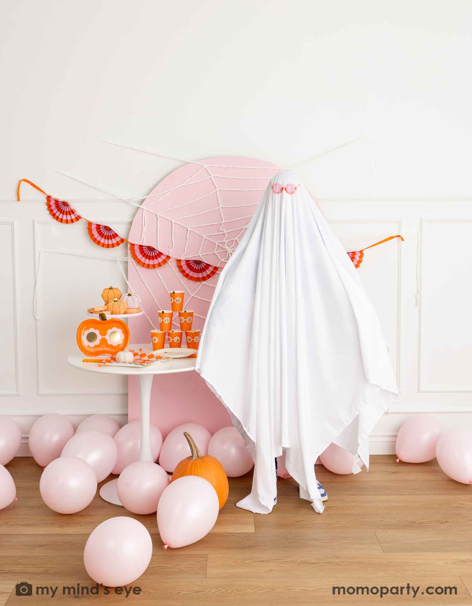 A kid's friendly Halloween party setup featuring a pale pink wooden curved backdrop board decorated with spider web, a party table filled with groovy Halloween pumpkin party tablewares from Momopartys and a bunch of pink balloons on the floor. A kid dresses as a sheet ghost wearing a pink sunglasses, making it groovy and fun vibe for this Halloween celebration.