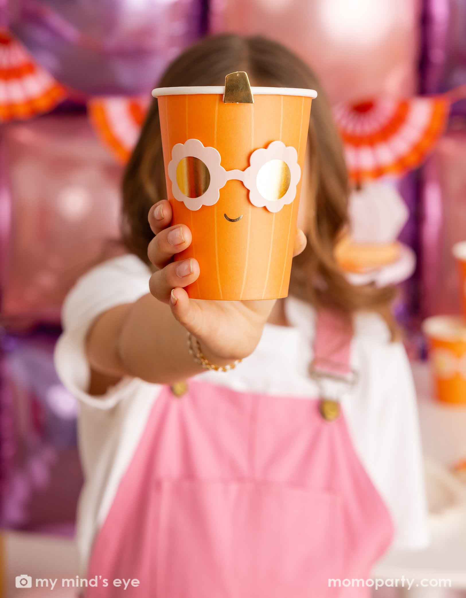 A girl in a pink overalls holding an orange pumpkin shaped paper party cup with a pair of daisy-shaped sunglasses from Momopartys in a festive Halloween setting featuring a balloon wall and an orange and pink bunting banner.