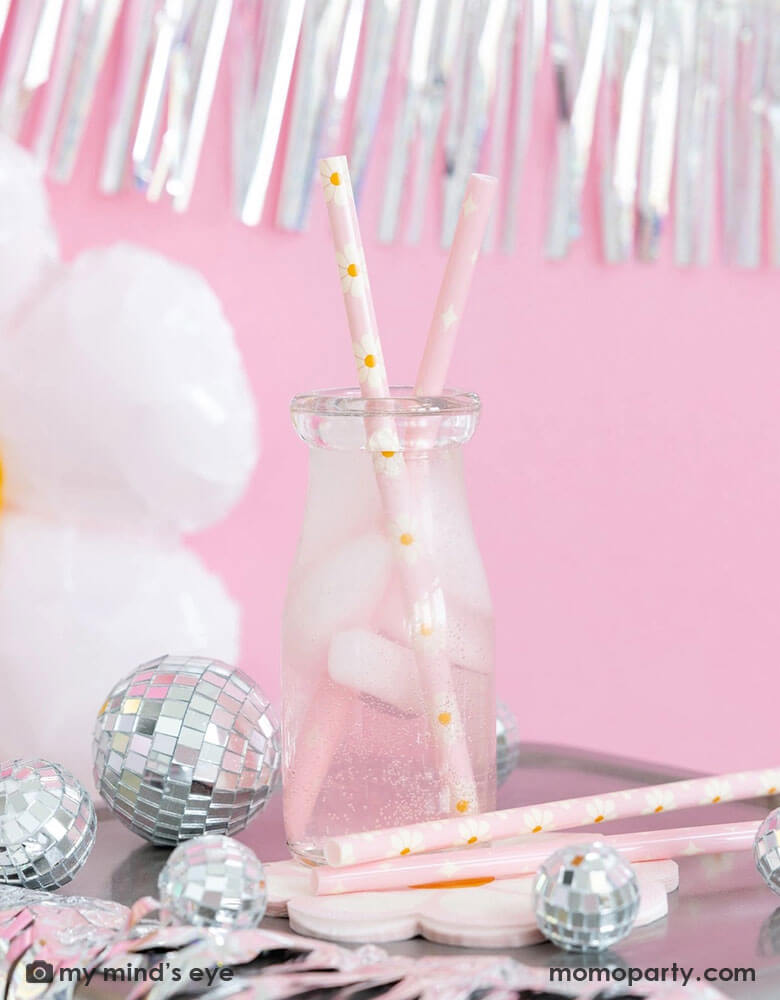 A milk bottle with pink soda and Momopartys's disco daisy reusable straws. Around the bottle there are Momopartys's disco daisy napkins and silver disco ball decorations. Above the the tableware is silver tinsel fringe garland, perfect for a groovy retro celebration!