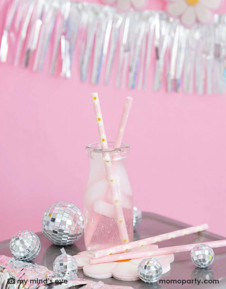 A milk bottle with pink soda and Momopartys's disco daisy reusable straws. Around the bottle there are Momopartys's disco daisy napkins and silver disco ball decorations. Above the the tableware is silver tinsel fringe garland, perfect for a groovy retro celebration!