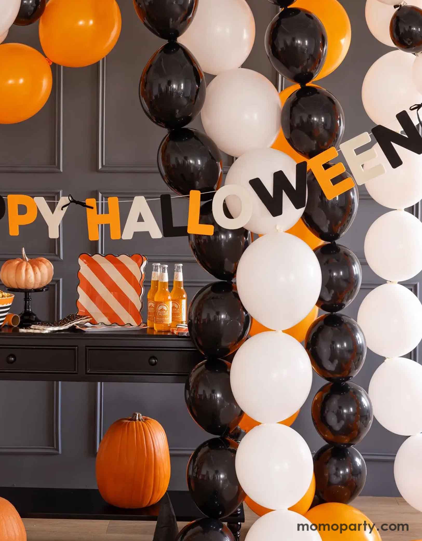 A Halloween-themed party setup with balloons in the classic Halloween colors of black, white and orange, along with Momopartys's Happy Halloween felt banner decoration, and multiple Halloween themed tablewares including plates, napkins, serving bowls and more.