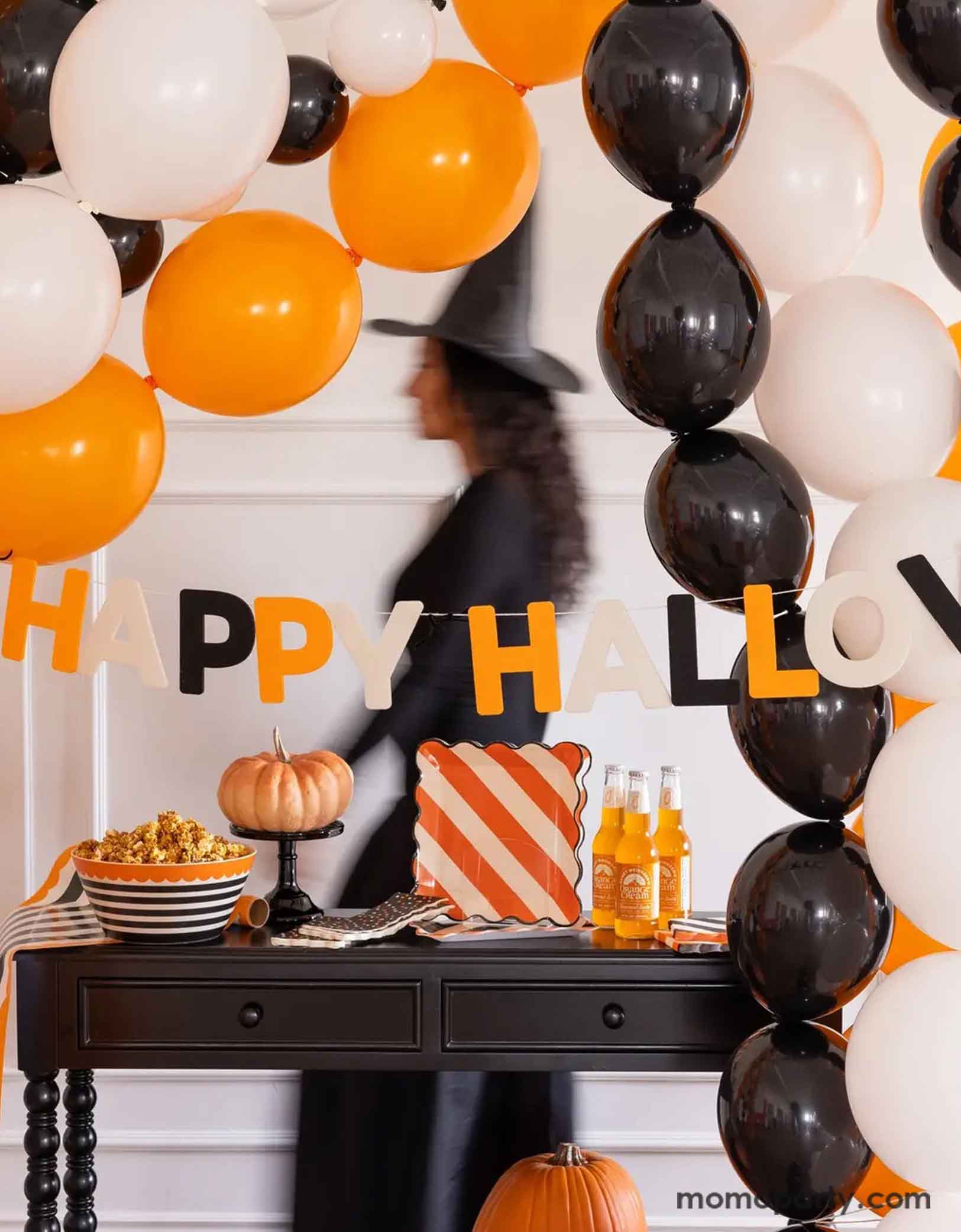 A Halloween-themed party setup with balloons in the classic Halloween colors of black, white and orange, along with Momopartys's Happy Halloween felt banner decoration, and multiple Halloween themed tablewares including plates, napkins, serving bowls and more. A lady in a witch costume walking by in motion in the back, making this a spooky yet fun inspo for a Halloween celebration.