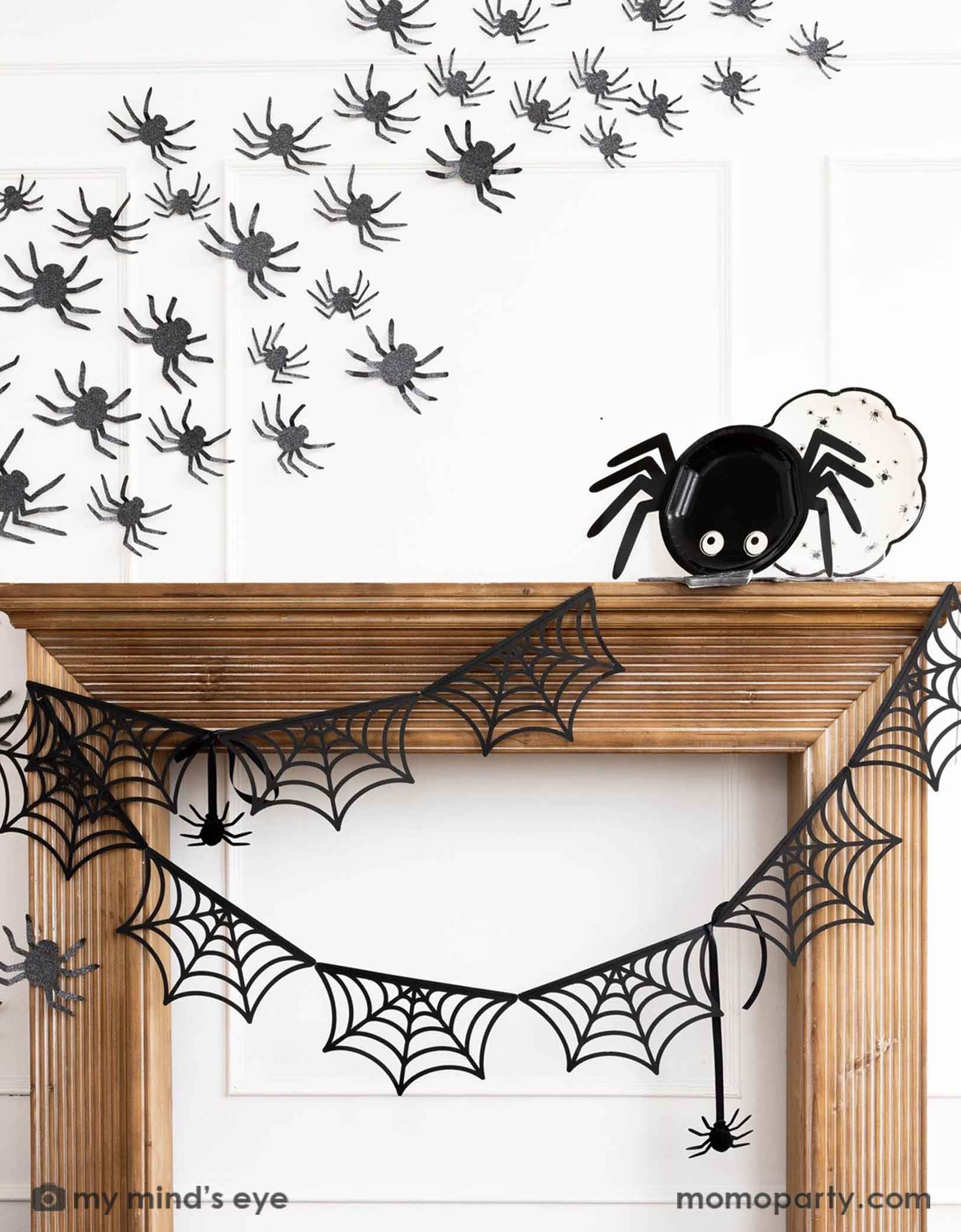 A wooden mantel against a white wall decorated with Halloween decorations from Momo  Party including the spider web banner, black spider decorations on the wall and the 14" x 7" spider shaped plate and a round plate with spider designs on it. Making this a kid's friendly cute Halloween decorating idea for the spooky season. 