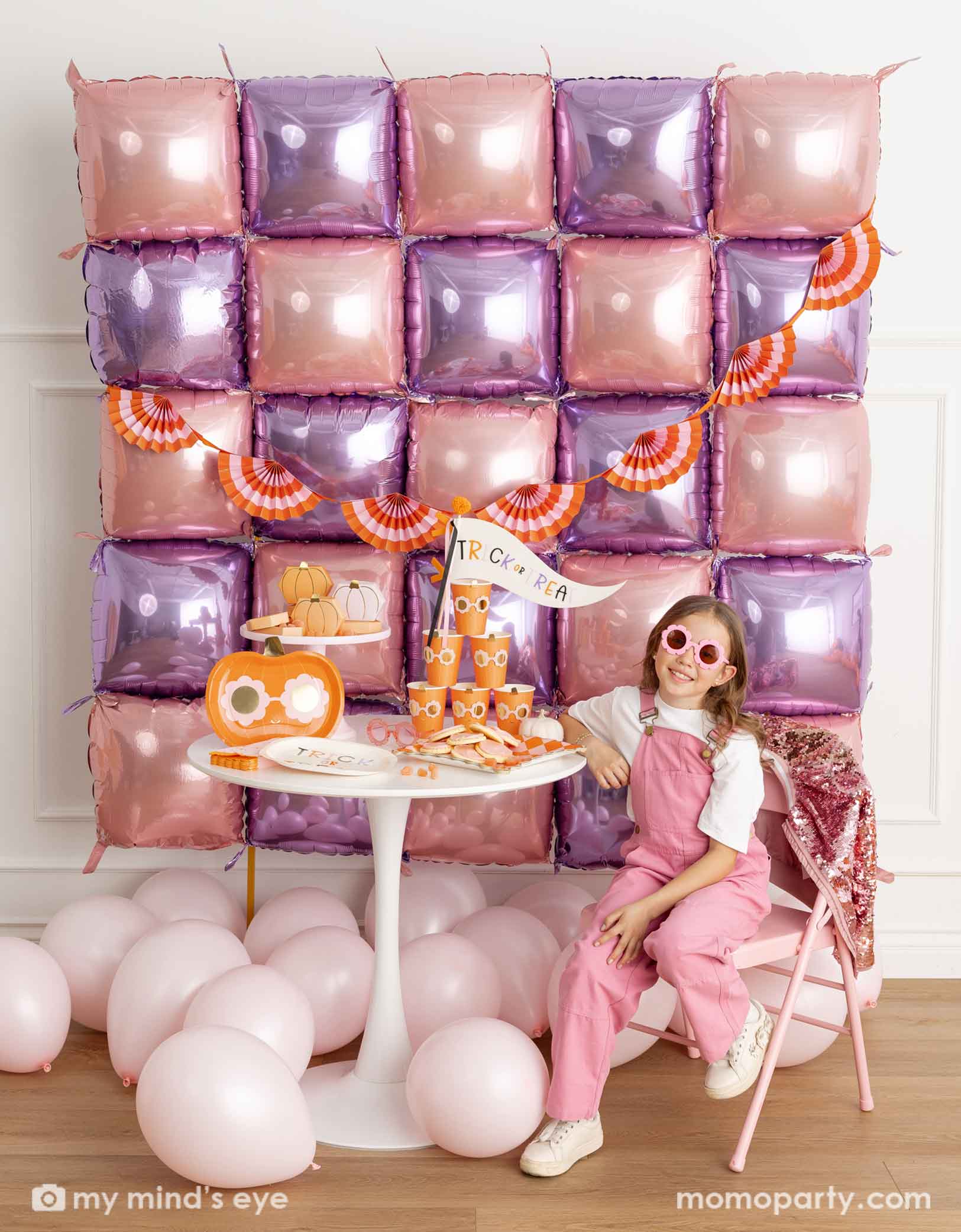 A girl in a pink overalls and daisy shaped sunglasses sitting in front of a decorative wall with pink and purple foil balloons adorned with an orange and pink bunting garland. Next to her is a white round table displaying groovy halloween pumpkin party tablewares including party plates, cups, napkins and the trick or treat felt party pennant. On the floor are some pale pink latex balloon, making this a festive kid's friendly pink Halloween celebration. 