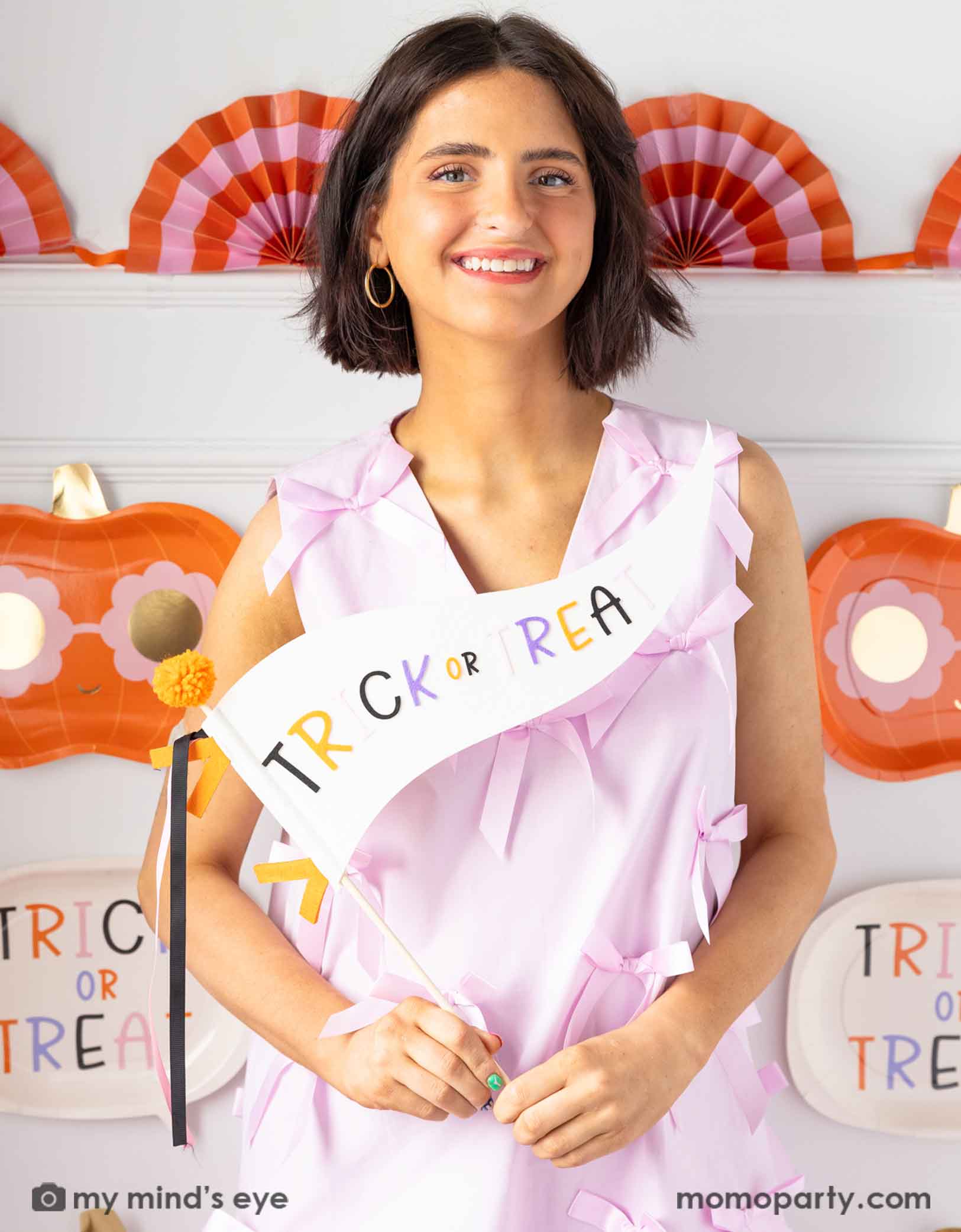 A lady in a pink dress holding Momopartys's 14" 'Trick or Treat' felt party pennant smiling into the camera with Halloween decorations on the wall in the background.