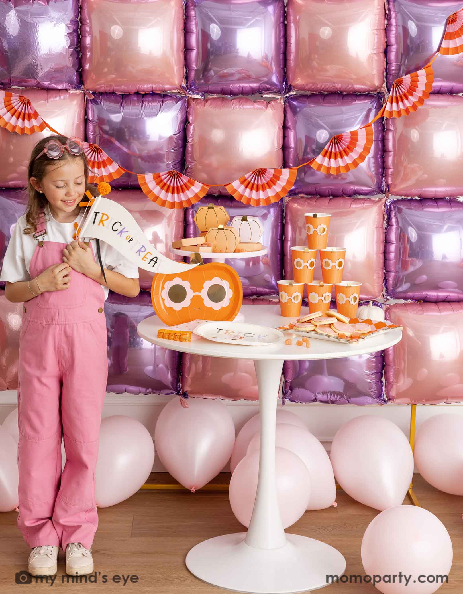 A girl in her pink overalls holding Momopartys's trick or treat felt party pennant standing in front of a decorative wall with pink and purple foil balloons adorned with an orange and pink bunting garland. Next to her is a white round table displaying groovy halloween pumpkin party tablewares including party plates, cups, napkins and treat boxes. On the floor are some pale pink latex balloon, making this a festive kid's friendly pink Halloween celebration. 