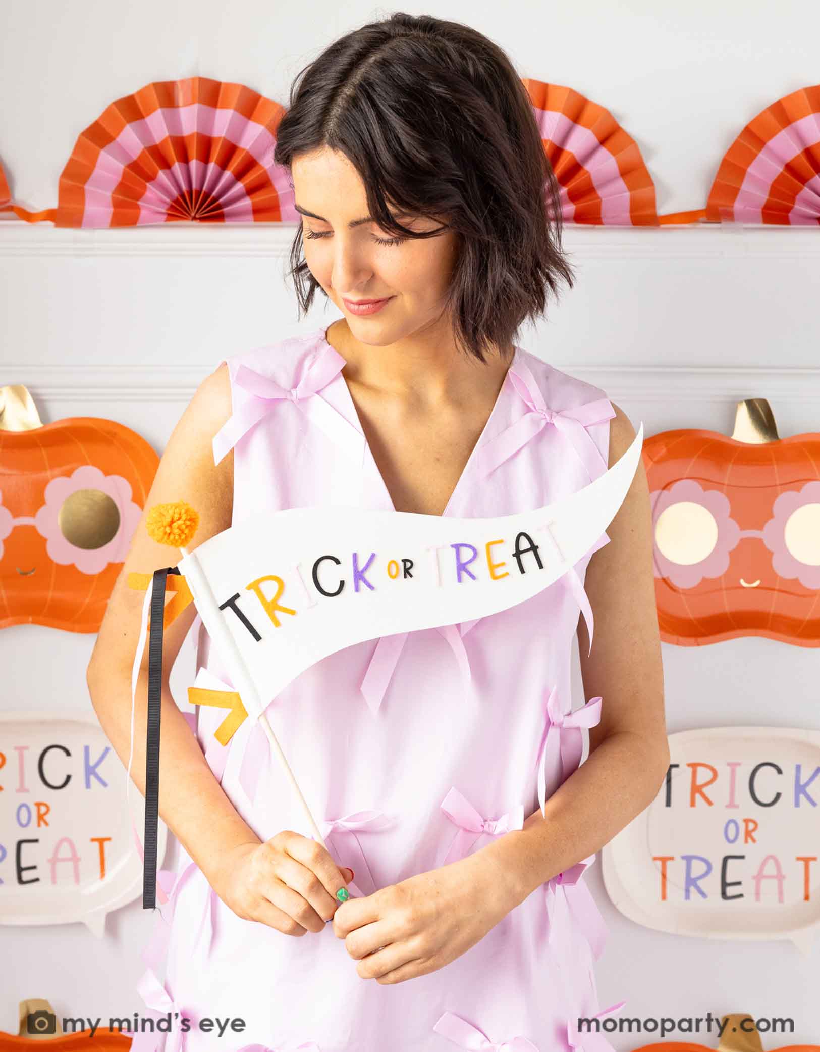 A lady in a pink dress holding Momopartys's 14" 'Trick or Treat' felt party pennant with Halloween decorations on the wall in the background.