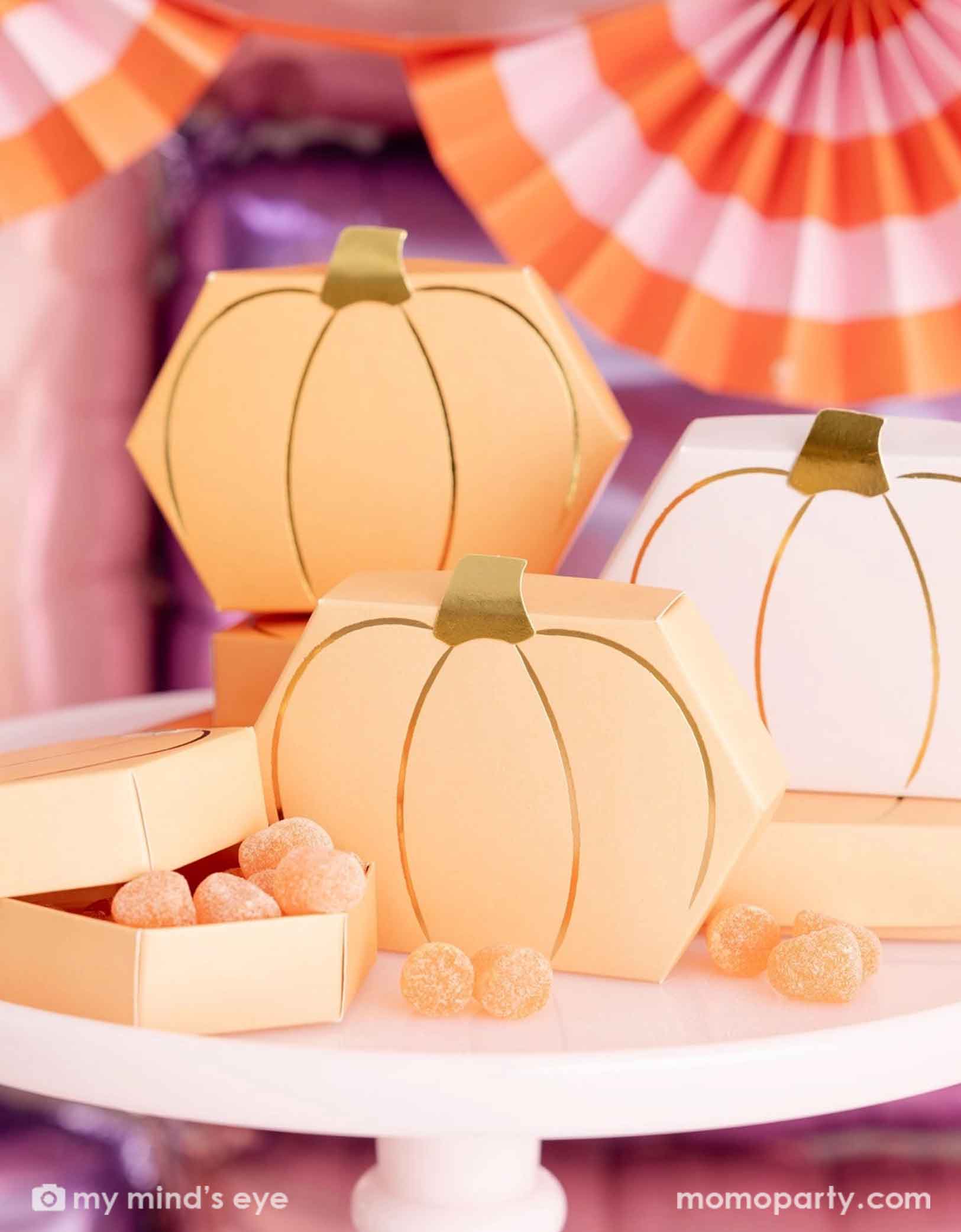 A close up of Momopartys's Pumpkin-shaped boxes by My Mind's Eye filled with orange gummies on a pale pink cake stand with festive pink Halloween decorative background with foil balloon wall and bunting garland.