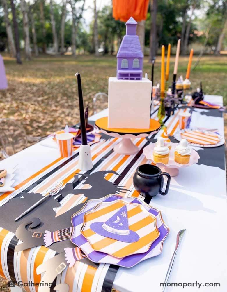 A Halloween witch themed party set up in a park featuring Momopartys's witch themed party supplies including purple and orange striped dinner plates, side plates and witch hat shaped napkins in the colors of purple, orange and peach. In the middle there's a haunted house shaped cake, some black and orange candles on ghost shaped candle stands on a black and orange striped table runner. Making this a great insp for kid's not-so-spooky Halloween party.