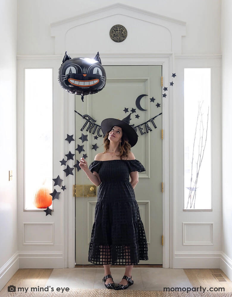 A lady dressed as a Halloween witch holding Momopartys's 24" black cat shaped foil balloon at the doorway decorated with Momopartys's 4ft Trick or Treat felt banner and glittered stars and moon wall paper decorations by My Mind's Eye. Behind the door through the side windows you can see a pumpkin and some spooky branches as the decorations, making this a perfect Halloween home decor idea for this spooky season!