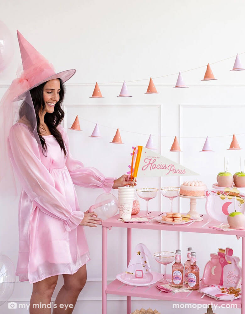 A pink Halloween party set up with a lady in her pink outfit and pink witch's hat. There's a pink cart filled with Momopartys's Hocus Pocus pink witch party supplies including the Hocus Pocus felt party pennant, pink party cups, pink cauldron shaped plates, pink witch hat shaped plates and pink potion shaped plates. On the wall there's the 3D pink witch banner hanging, along with the pink cakes, treats, snacks and drinks on the cart, it's perfect for a girly Halloween celebration!