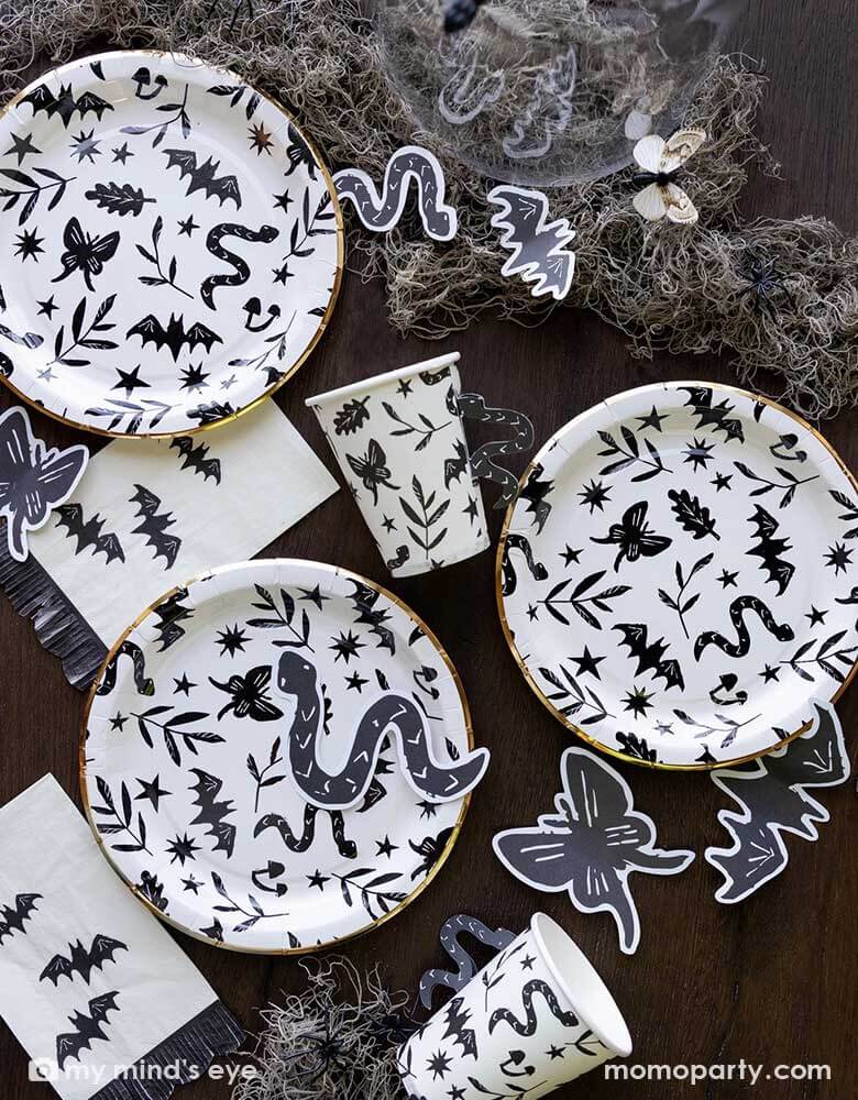 A table covered with Momopartys's spooky icon party supplies by My Mind's Eye including paper plates, cups, napkins and paper confetti. with some spooky dry grass around, this eerie line is perfect for a stylish Halloween party this spooky season.