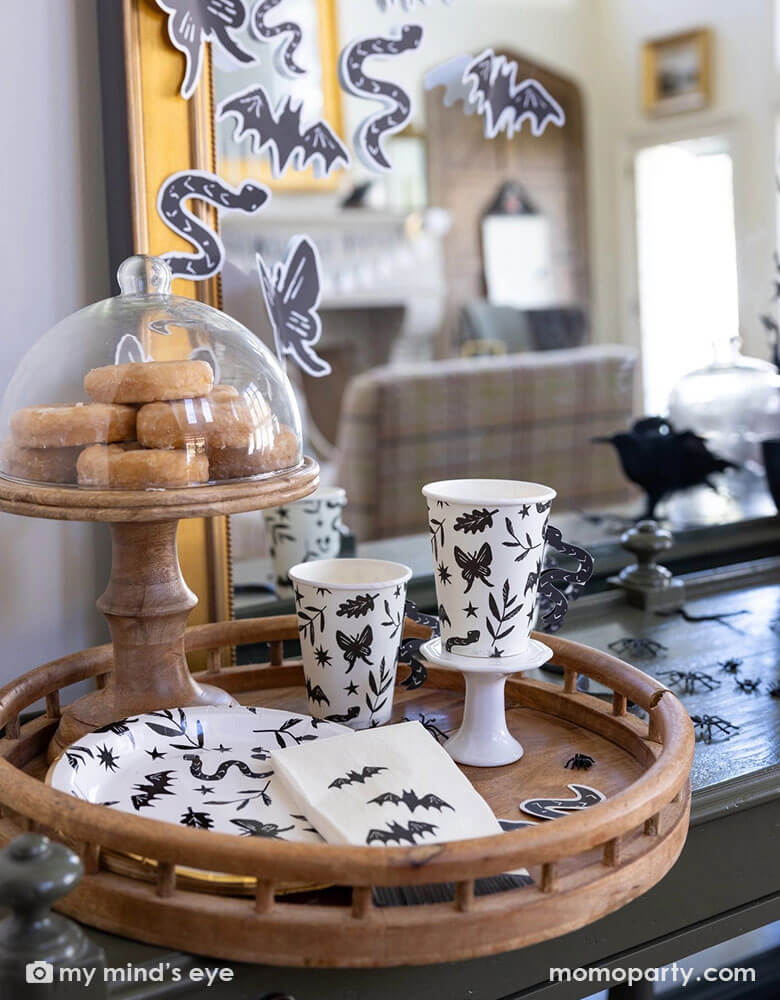 A spooky yet stylish Halloween table decoration featuring Momopartys's spooky icon party supplies collection including the snake handle party cups, the black and white spooky icon round plates and the bat guest napkins in a wooden tray which has some glazed donuts on a wooden cupcake stand. In the back there's a large mirror covered with Halloween paper decorations in black and white of Halloween elements including snakes, butterflies, bats, and butterflies. Making this a perfect stylish Halloween inspo.
