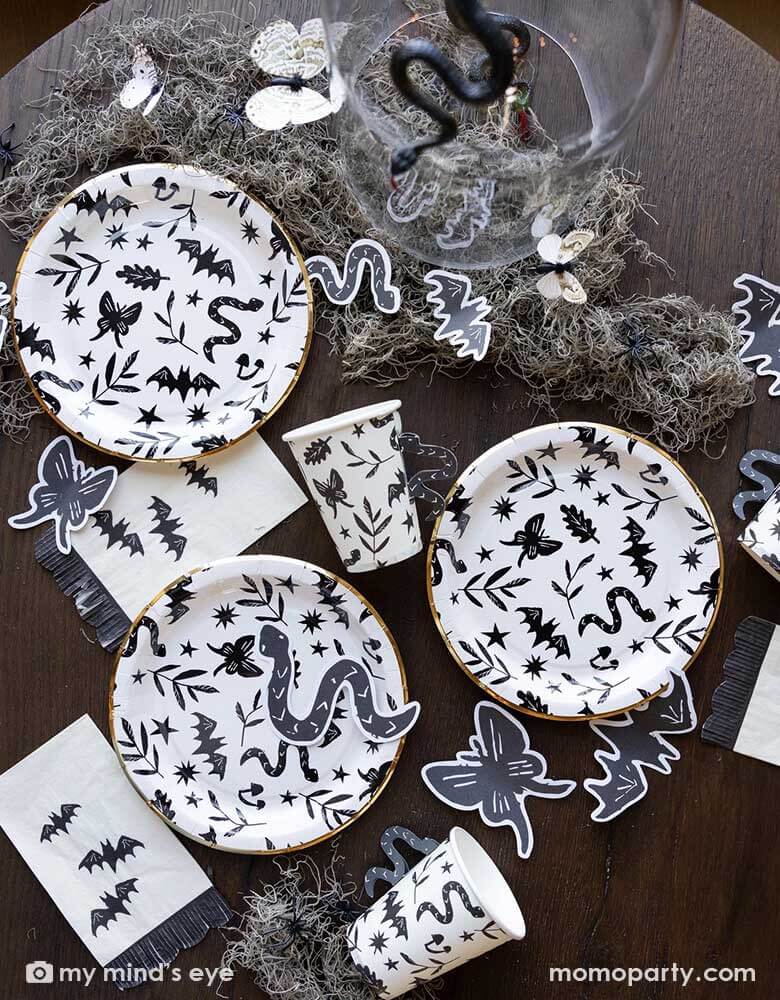 A table covered with Momopartys's spooky icon party supplies by My Mind's Eye including paper plates, cups, napkins and paper confetti. with some spooky dry grass around, this eerie line is perfect for a stylish Halloween party this spooky season.