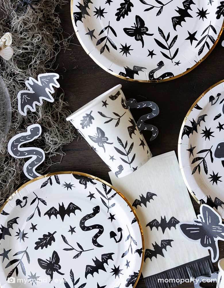 A table covered with Momopartys's spooky icon party supplies by My Mind's Eye including paper plates, cups, napkins and paper confetti. with some spooky dry grass around, this eerie line is perfect for a stylish Halloween party this spooky season.