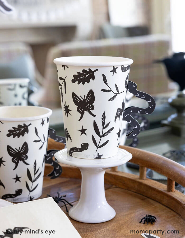 A close up shot of Momopartys's 12 oz Snake Handle Party Cups by My Mind's Eye on a wooden table covered with spider decoration and confetti. These spooky cups not only come in a fun snake handle design, but they will also make your party stand out in a unique way. Don't settle for boring party cups, add a touch of fun with these cups!