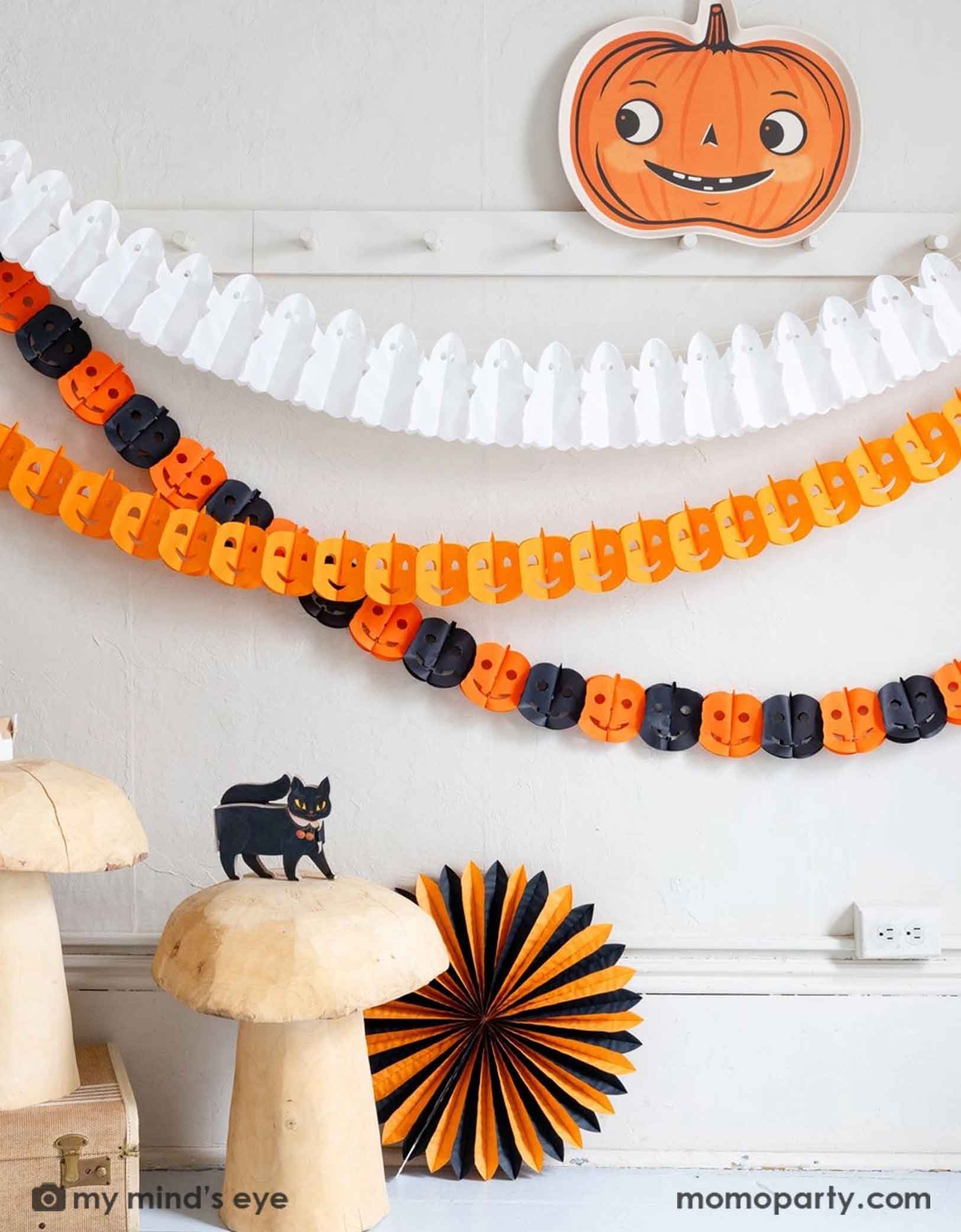 A white wall with coat hanging rack decorated with Halloween-themed decorations including Momopartys's pumpkin-shaped bamboo tray, ghost tissue garland, orange Jack-o'-lantern tissue garland, plus an orange and black Jack-o'-lantern tissue garland hung on the wall. Underneath is a cat figurine on a bunch of wooden mushroom stools which are next to a orange and black tissue paper fan decoration. A simple and modern way to decorate your home in the Halloween spooky season.
