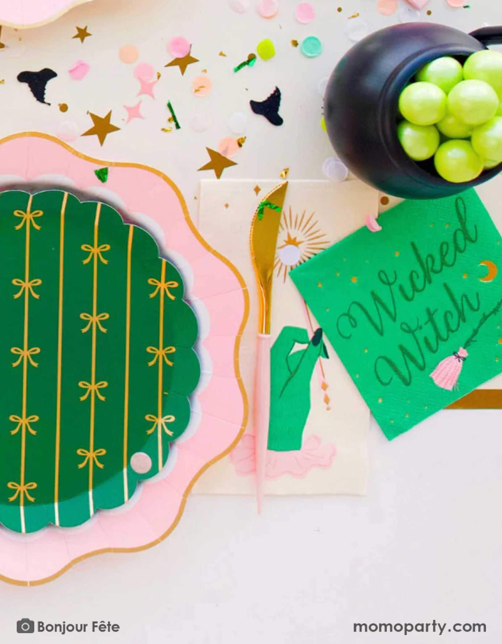 A whimsical Wicked themed party table setting featuring multiple Wicked themed party supplies from Momopartys including the Wicked Witch 5" small napkins, the magic wand guest napkins, a large pink dinner plate and an emerald green side plates with gold foil bows pattern, along with Wicked themed confetti spread out on the table and a mini cauldron filled with green grapes, it's a perfect party inspo for a fun and magical Wicked themed party in the Halloween spooky season.
