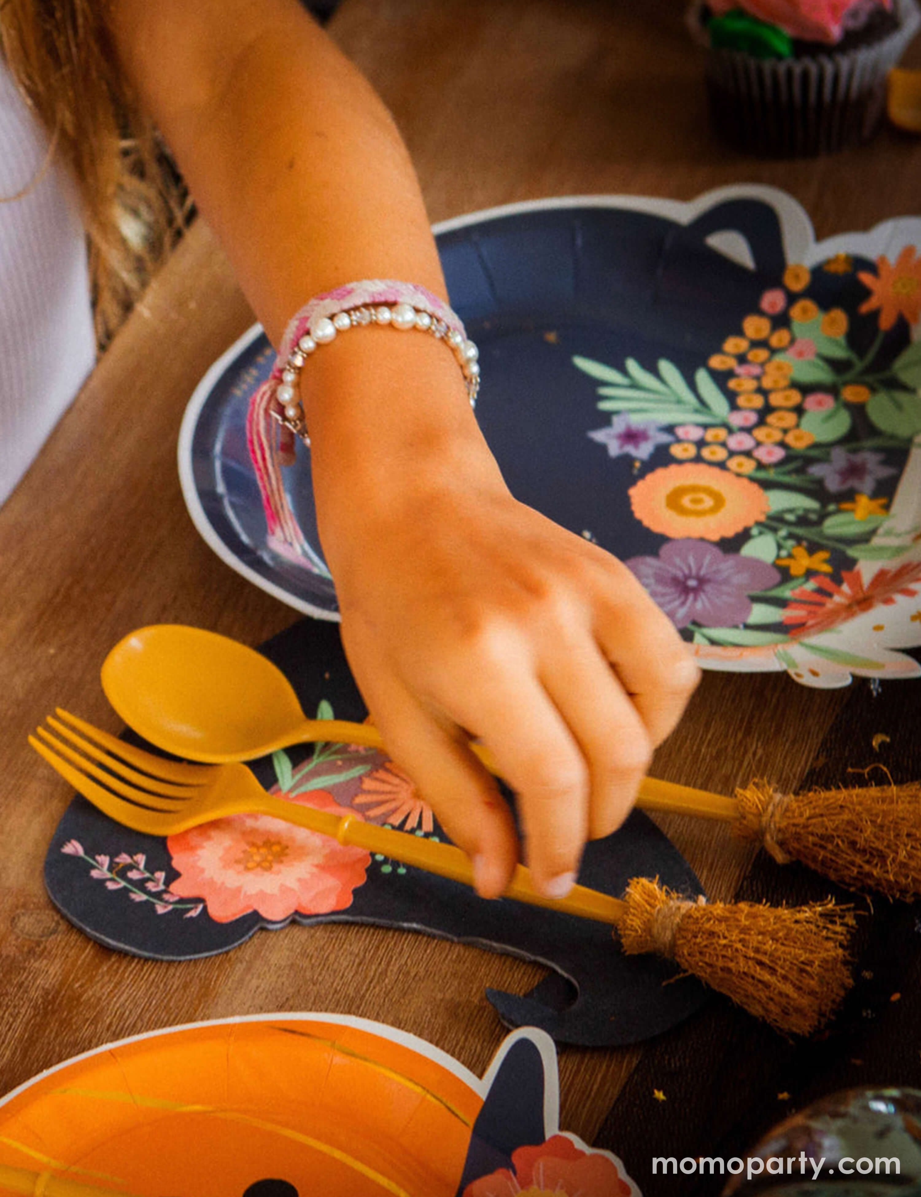 A girl's hand reaching Momopartys's witch's broom cutlery set on a witch themed Halloween party table featuring cauldron shaped party plates, pumpkin shaped party plates and witch hat shaped napkins with floral designs on them. Making this a great inspo for a boho and chic Halloween party tablescape. 