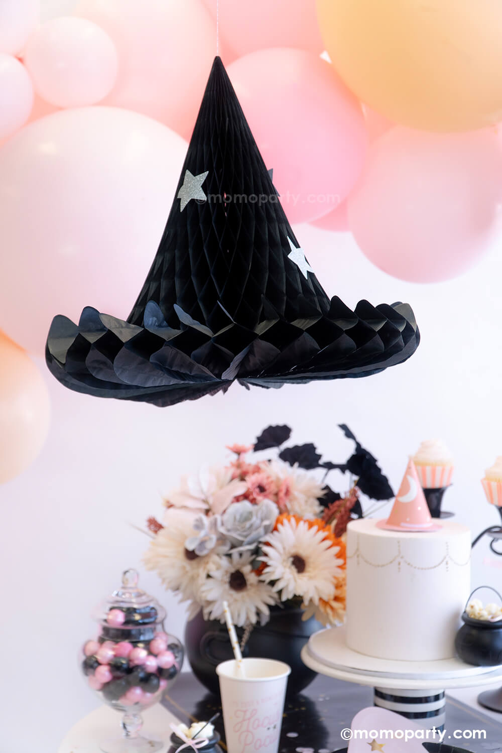 Momo Party's Hocus Pocus Witch Hat Honeycomb decoration hung next to an adorable pink balloon garland with various sized balloons in pastel pink, pink and blush colors. Below it is a pink inspired Halloween party table featuring witch themed party supplies from Momopartys including Hocus Pocus party cups, napkins, witch hat shaped plates and some pink and black themed treats. This makes a great inspo for a sweet girly pink Halloween that's perfect for this spooky season.
