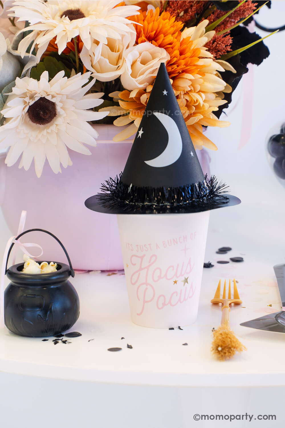 A witch themed Halloween party table styled by Momopartys featuring pink Halloween party supplies including the pink Hocus Pocus party cup which is topped with the witch party hat with a crescent moon on it. On the table there's a broom shaped fork next to a witch hat shaped plate and a mini cauldron filled with popcorn. With the fall flower arrangement in the pink cauldron in the back and black confetti around the table, it makes a spooky cute tablescape inspo for a girly Halloween bash this spooky season