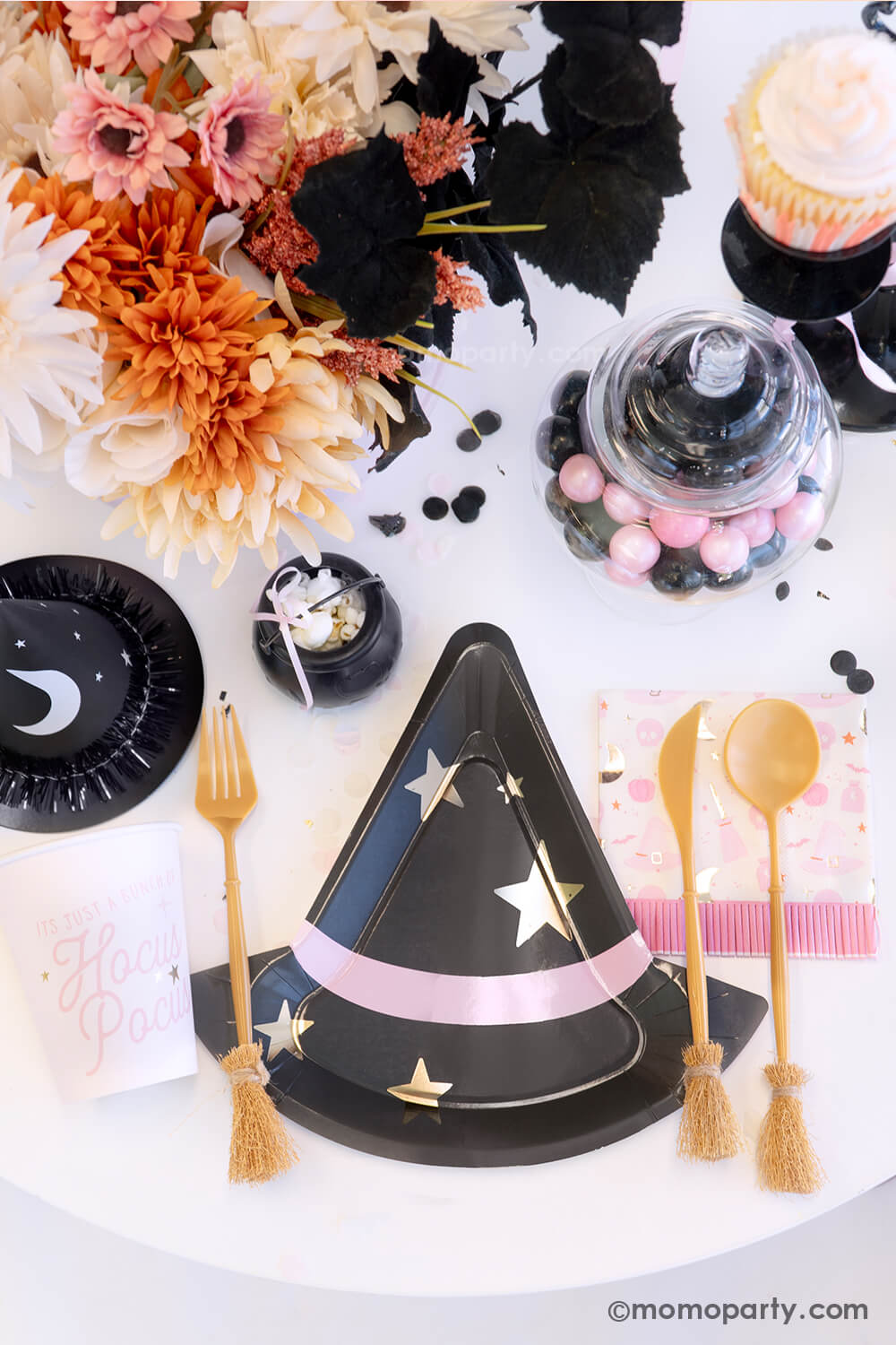 A witch themed Halloween party table styled by Momopartys featuring pink Halloween party supplies including the  pink witch hat shaped plate, witch's broom cutlery set, Hocus Pocus party cups and napkins, and the witch party hat. On the table, there's mini cauldron with popcorn, some pink and black gum balls and a fall flower arrangement in the pink cauldron - making it a spooky cute tablescape inspo for a girly Halloween bash this spooky season.