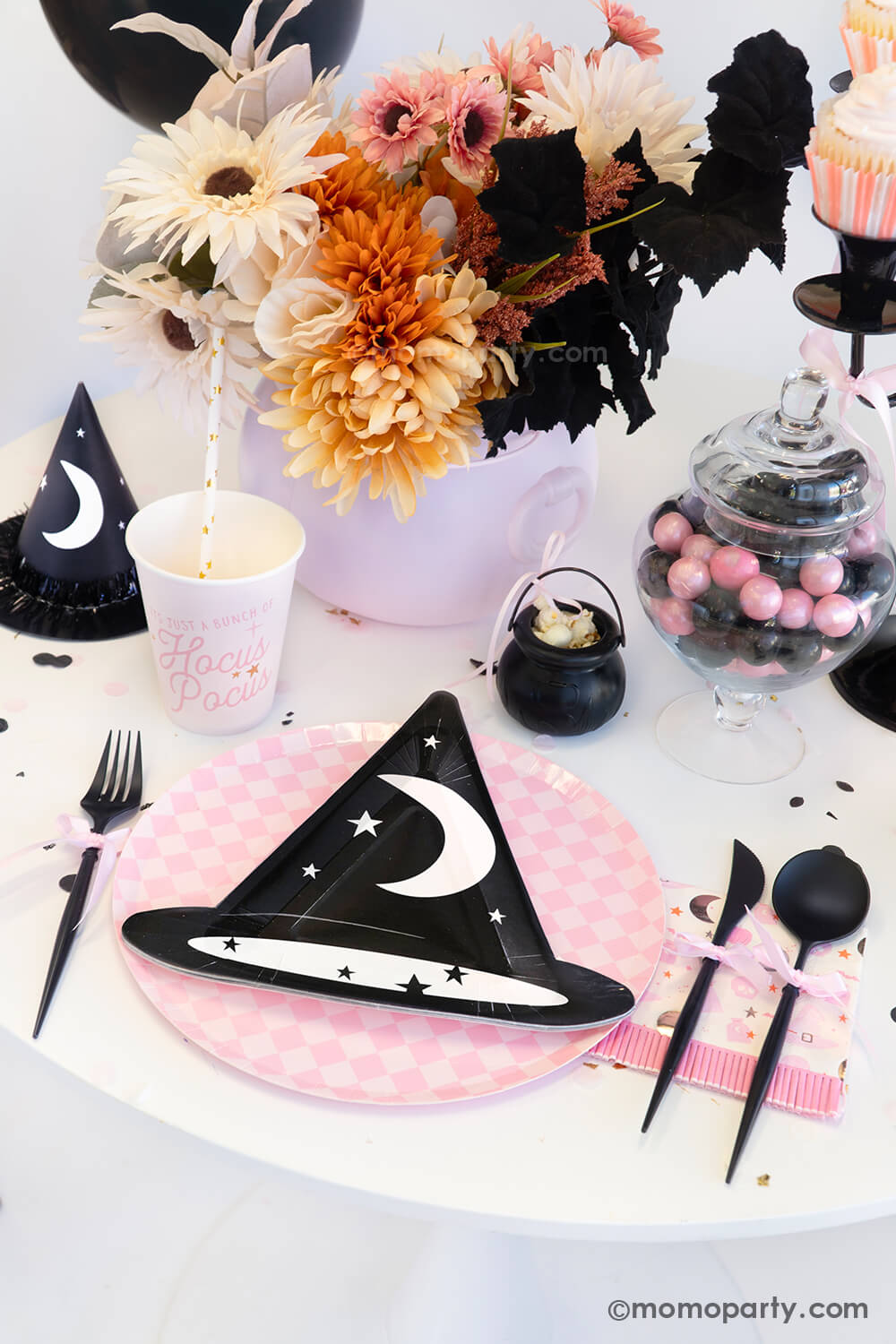 A witch themed Halloween party table styled by Momopartys featuring pink Halloween party supplies including the  pink Hocus Pocus party cup, napkin, witch hat shaped plate on a pink checkered plate, and a witch party hat. On the table, there's mini cauldron with popcorn, some pink and black gum balls and a fall flower arrangement in the pink cauldron - making it a spooky cute tablescape inspo for a girly Halloween bash this spooky season.