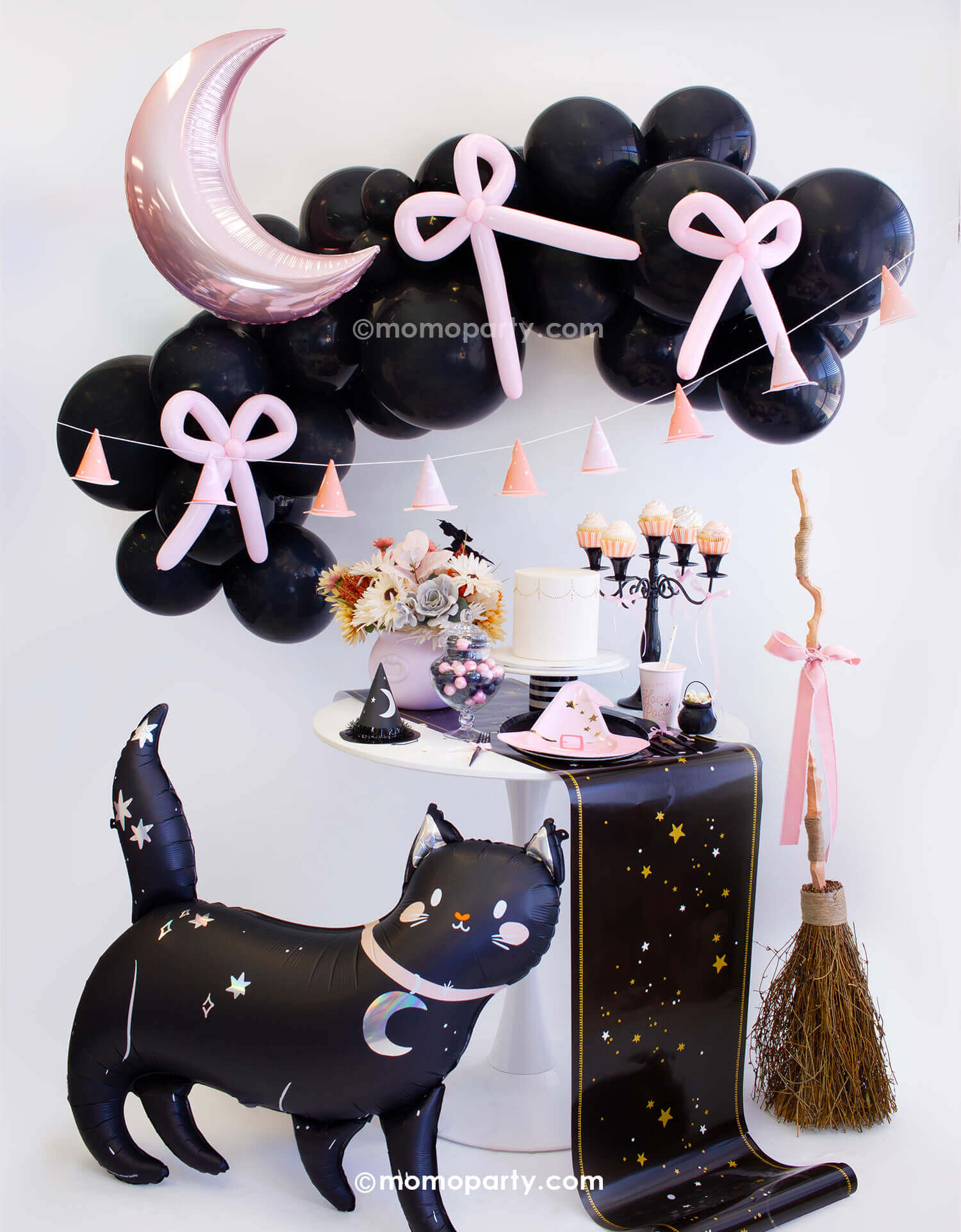 An adorable kid's friendly witch themed Halloween party look styled by Momopartys. With black balloon garland adorned with pastel pink balloon bows and the pink crescent moon shaped foil balloon, along with pink witch hat party banner as the backdrop. On the party table there are witch themed tableware including pink witch hat shaped plates, Hocus Pocus napkins and party cups, along with mini cauldrons, witch hats and treats. In the front there are a cute black cat foil balloon and a broom with a pink bow.